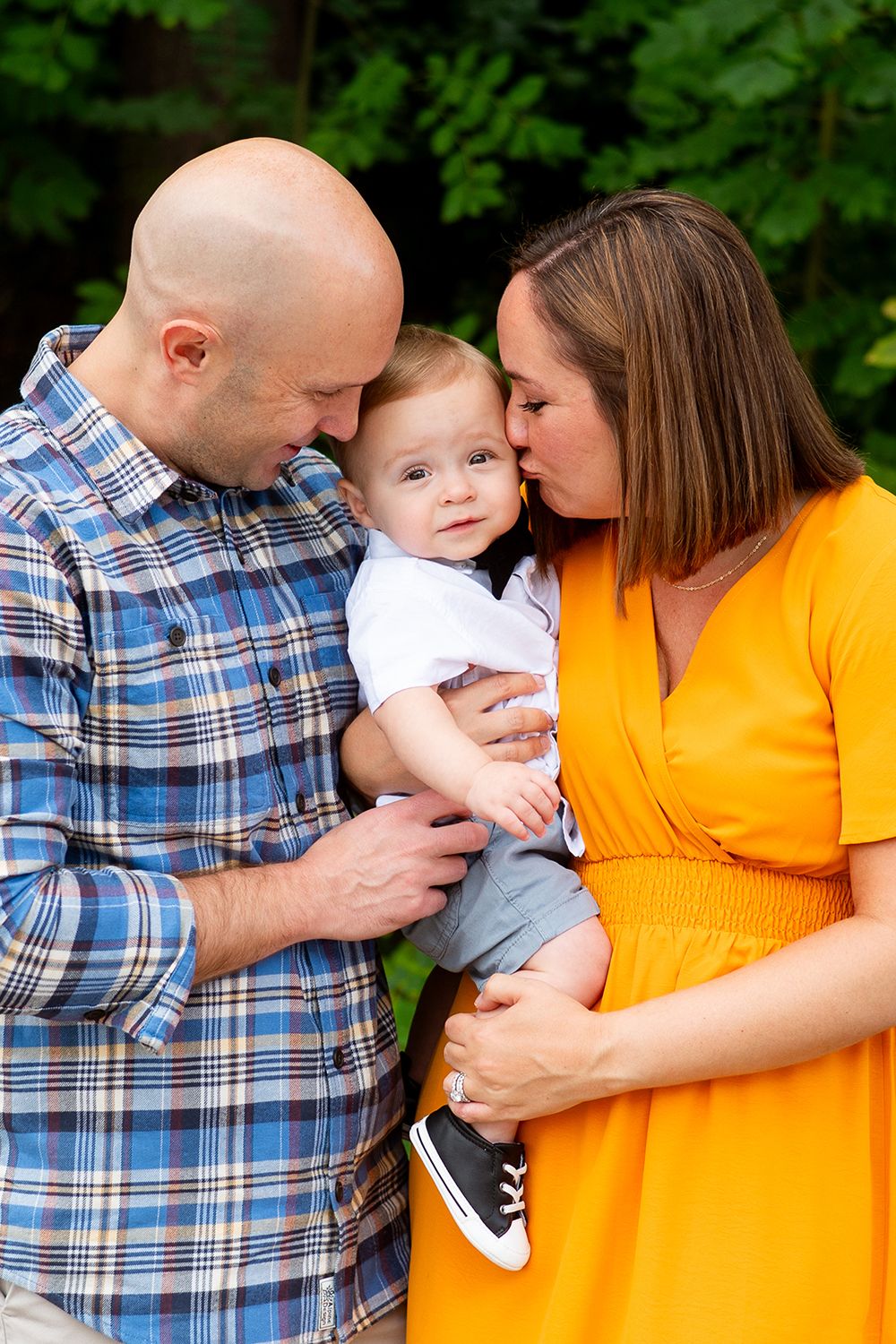 Intimate family portrait outdoors with baby wearing white onesie between parent in plaid shirt and yellow dress.