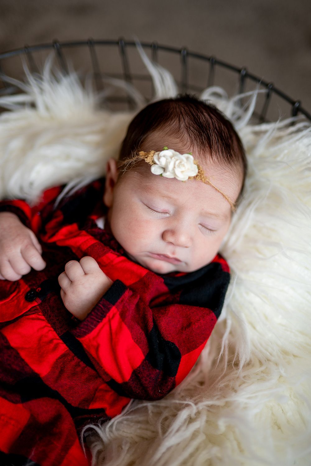 Sleeping baby wearing red buffalo plaid outfit rests on white fur with small floral headband.