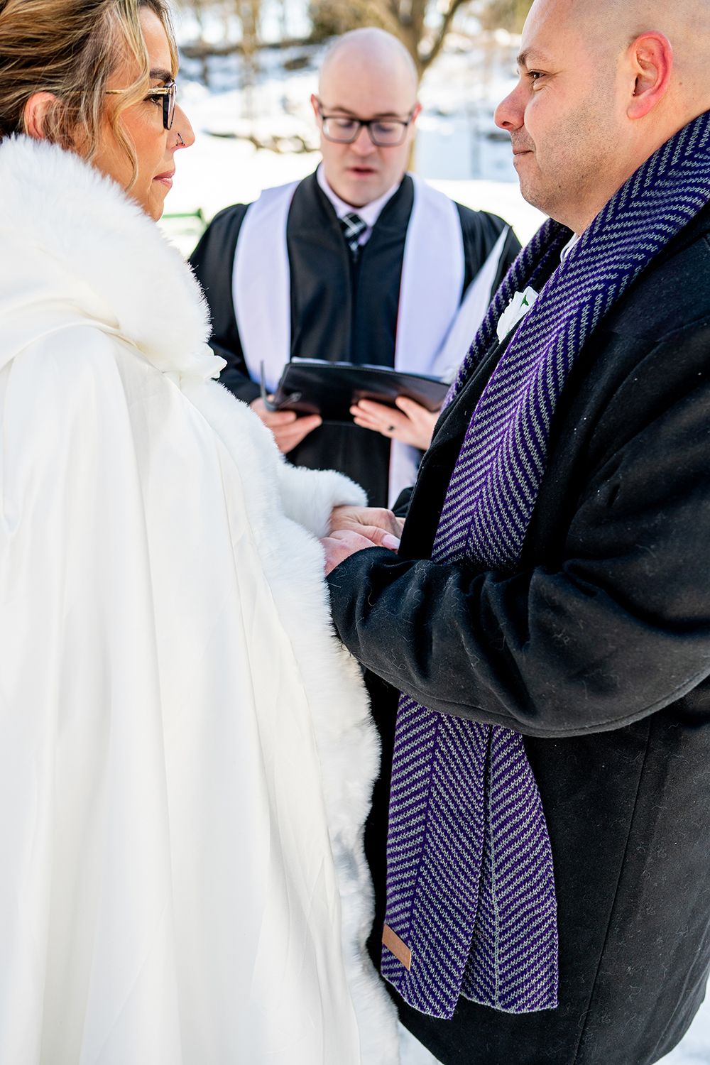 A winter wedding ceremony takes place outdoors with an officiant performing the service in the snow.