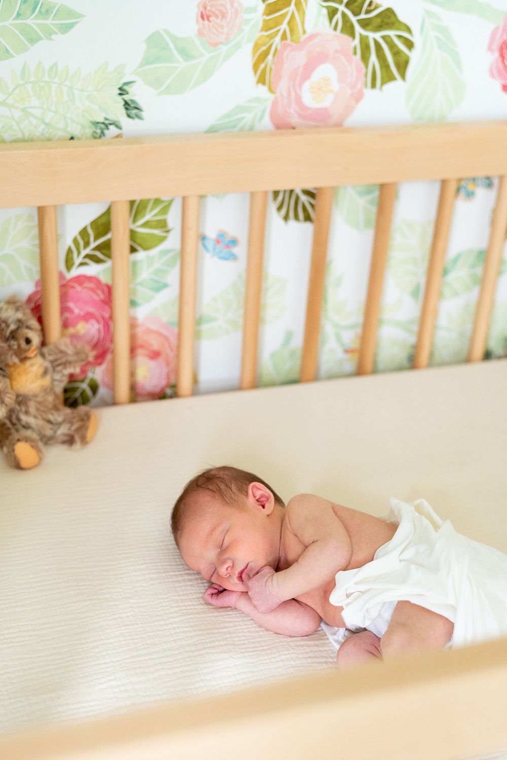 A sleeping newborn rests peacefully in a wooden crib with floral bedding and a plush teddy bear.