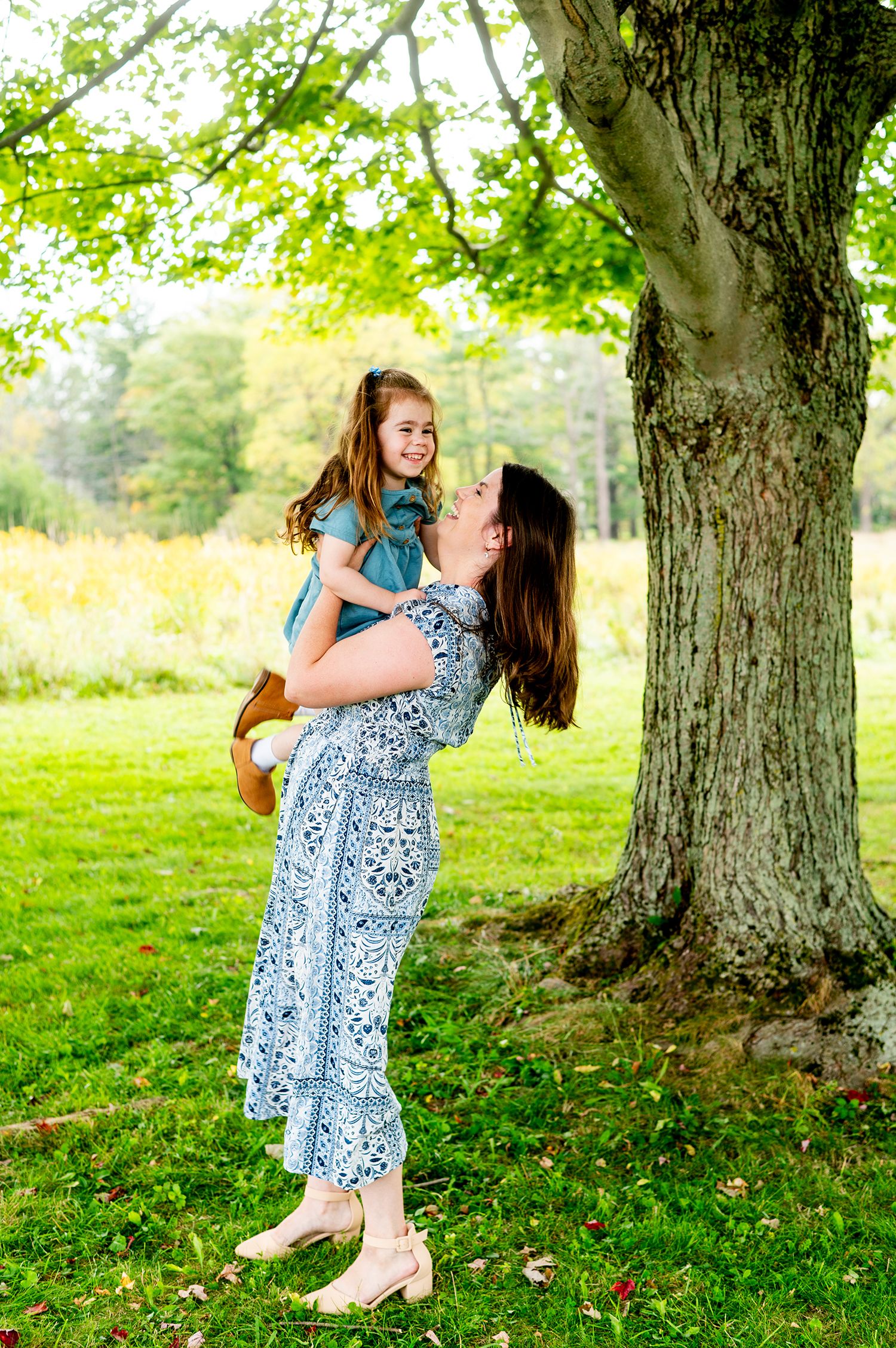 A mother in a floral dress lifts her smiling daughter outdoors near a large tree on a green lawn.