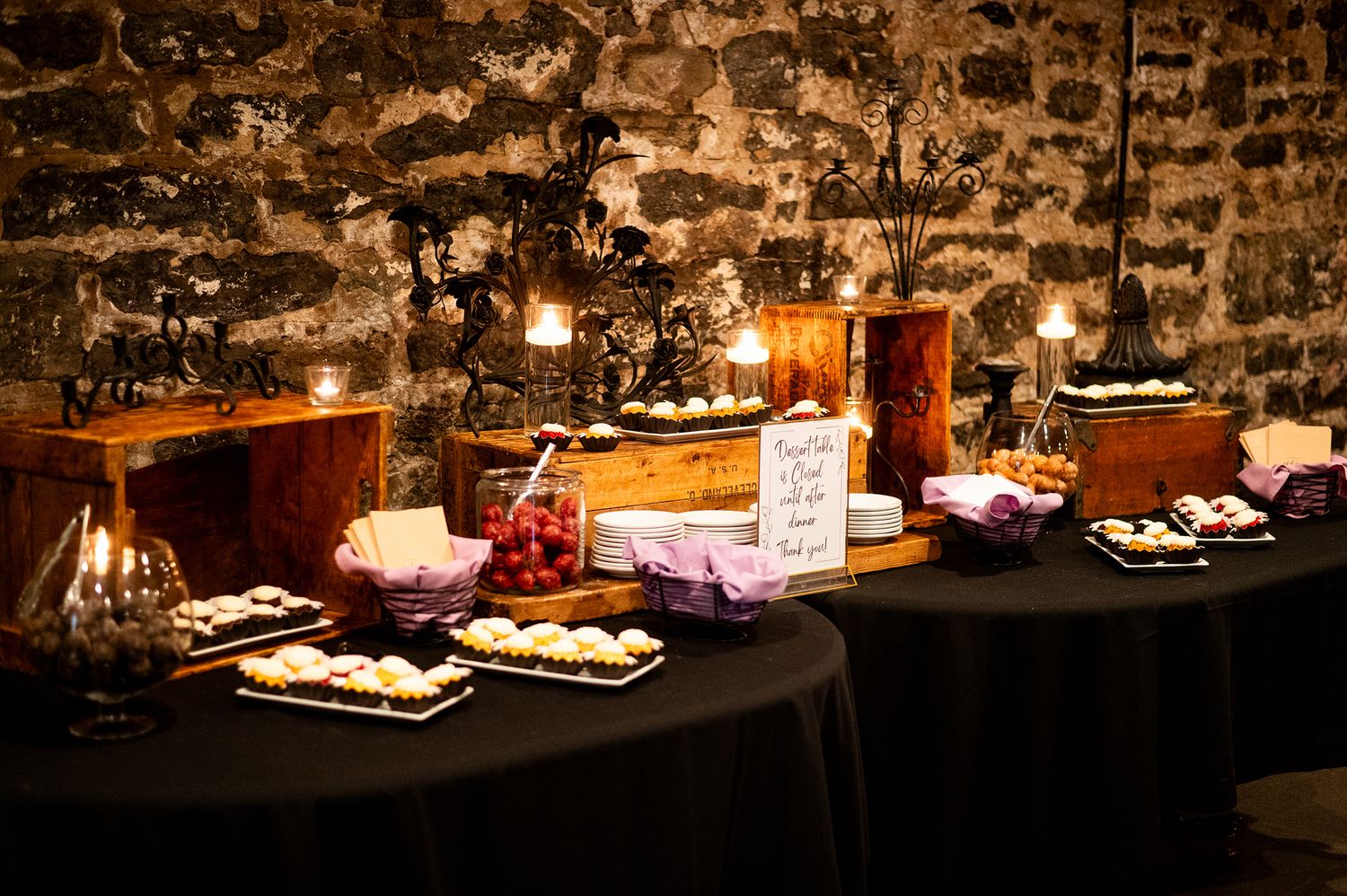 Elegant candlelit dessert buffet table with assorted sweets against rustic stone wall.