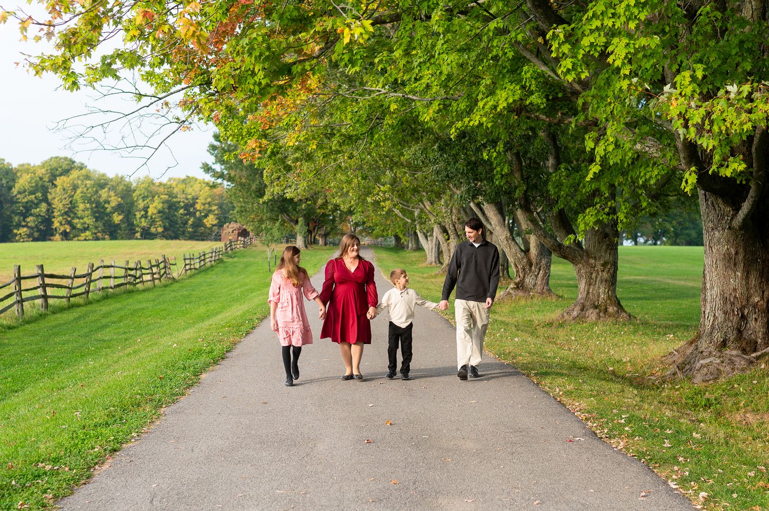 Family of four walking hand-in-hand down a tree-lined path on a sunny autumn day.