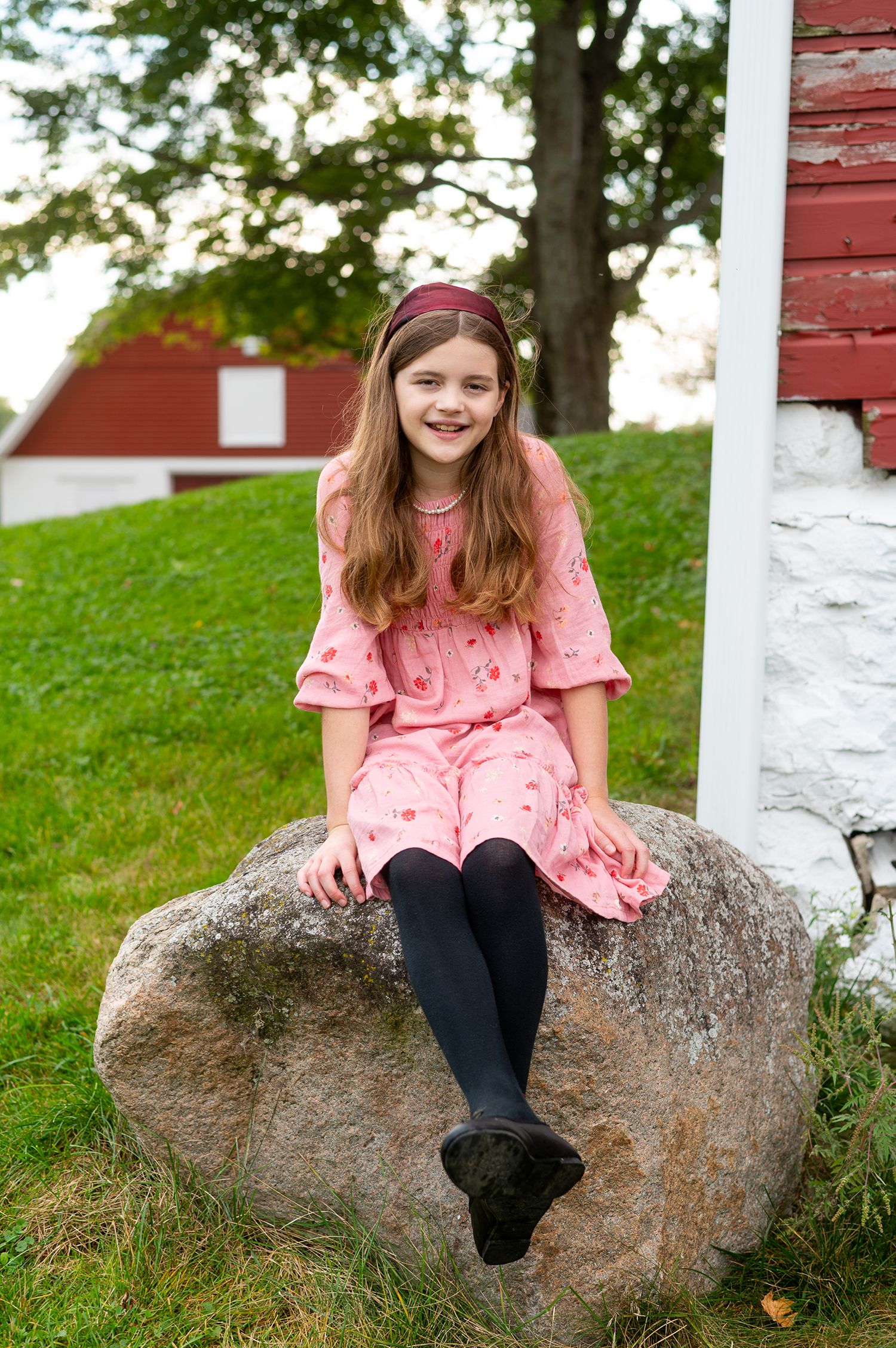 A smiling young girl in a pink dress and dark tights sits on a large rock near a red barn.
