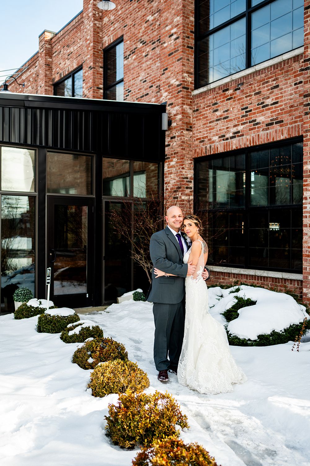 A couple poses for a winter wedding photo against a rustic brick building with snow-covered shrubs.