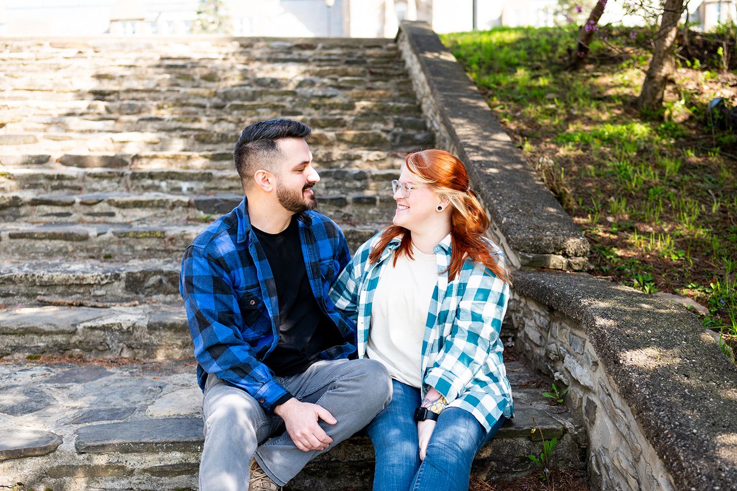 Two people sit close together on rustic stone steps surrounded by dappled sunlight and greenery.