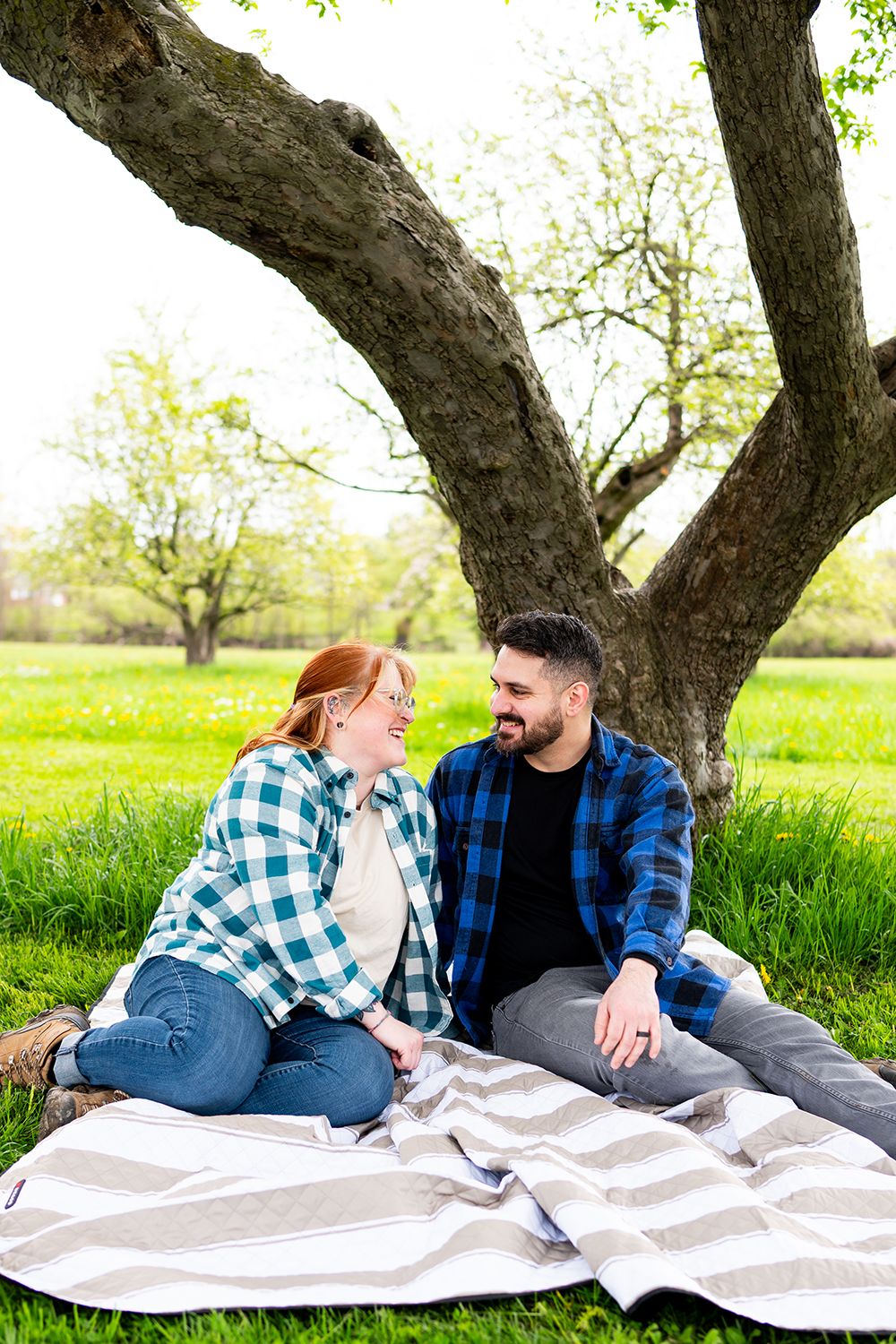 Couple in matching plaid shirts share a happy moment on a blanket under a large tree in a spring meadow.