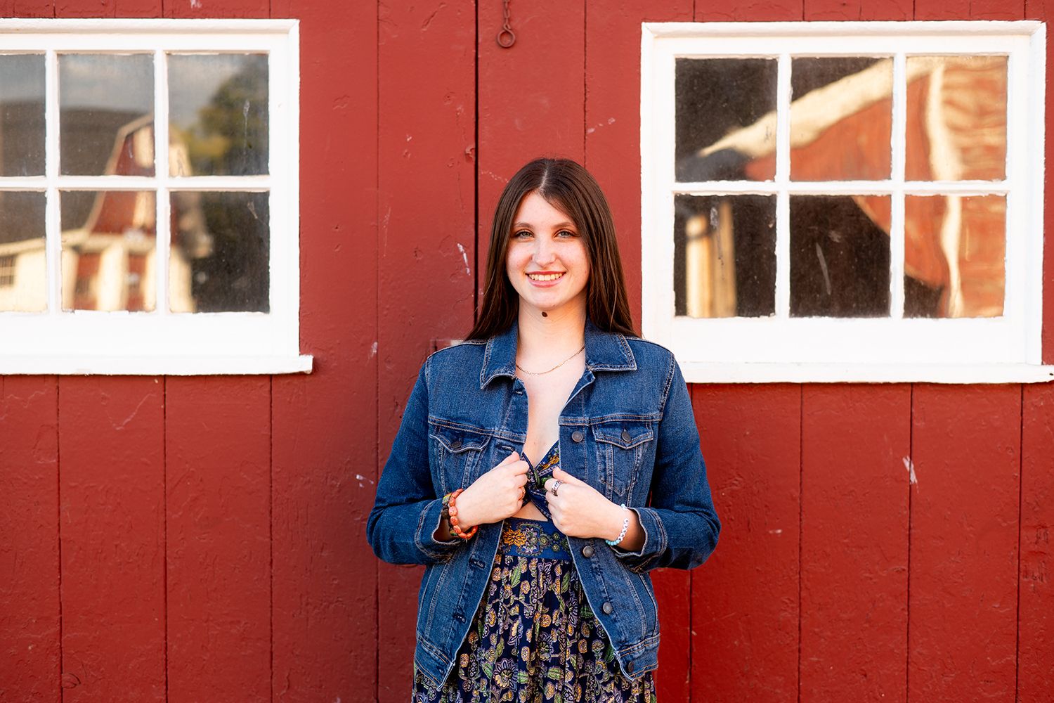 Someone wearing a denim jacket and floral dress stands in front of a red barn wall with white-framed windows.