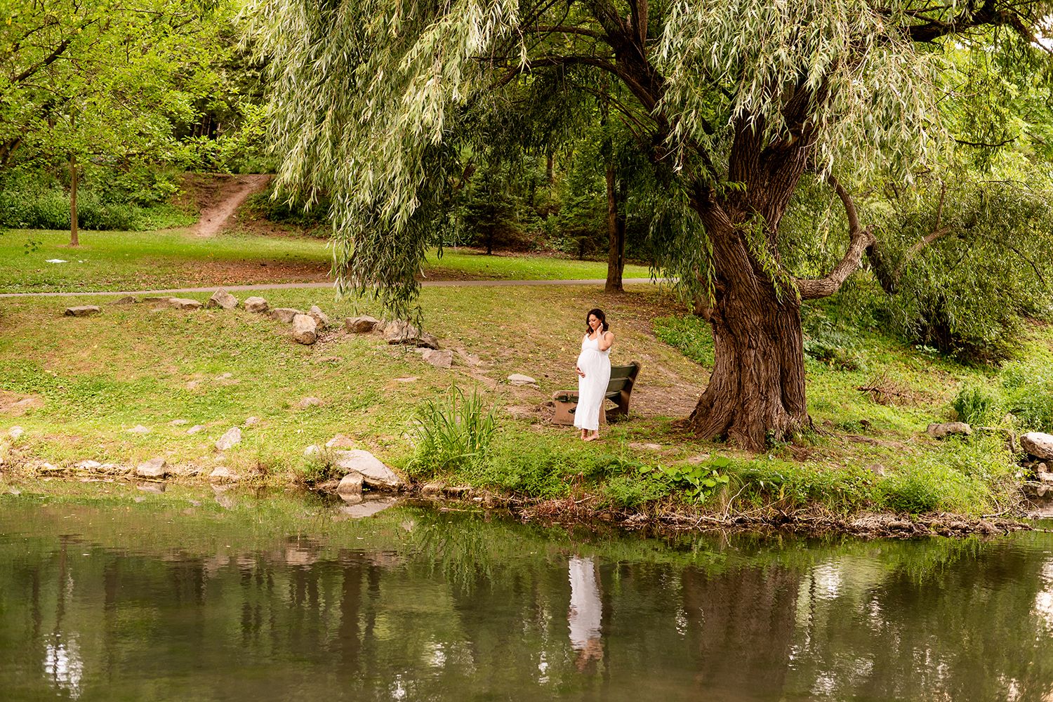 A person in white stands by a large willow tree next to a tranquil pond in a scenic park setting.