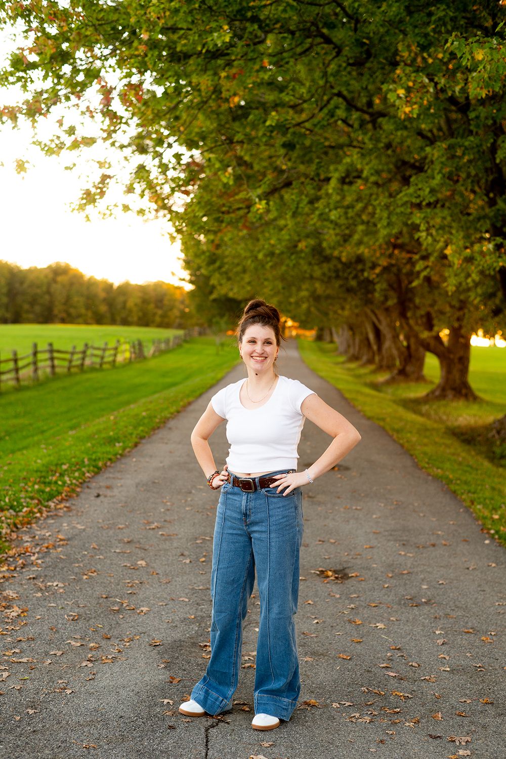 Person wearing jeans and white shirt poses on tree-lined path during golden autumn sunset.