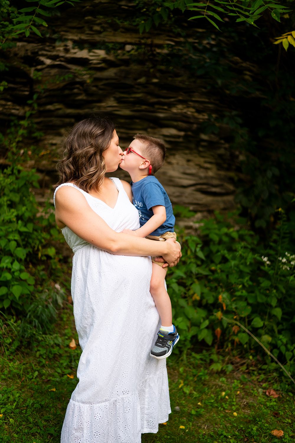 A tender moment captured outdoors as a white-dressed woman holds and kisses a small child against a rocky backdrop.