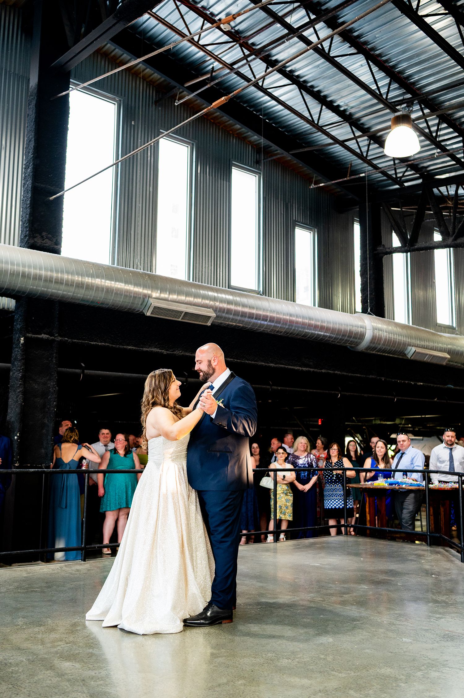 A couple shares their first dance in an industrial warehouse venue with wedding guests watching in the background.
