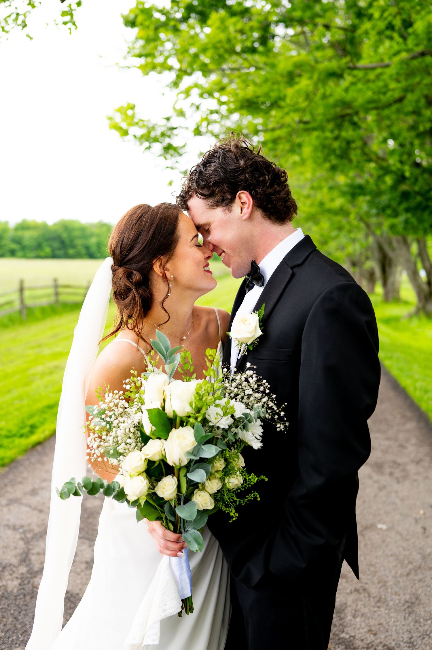 Romantic wedding couple shares intimate moment on tree-lined gravel path with white floral bouquet and spring greenery.