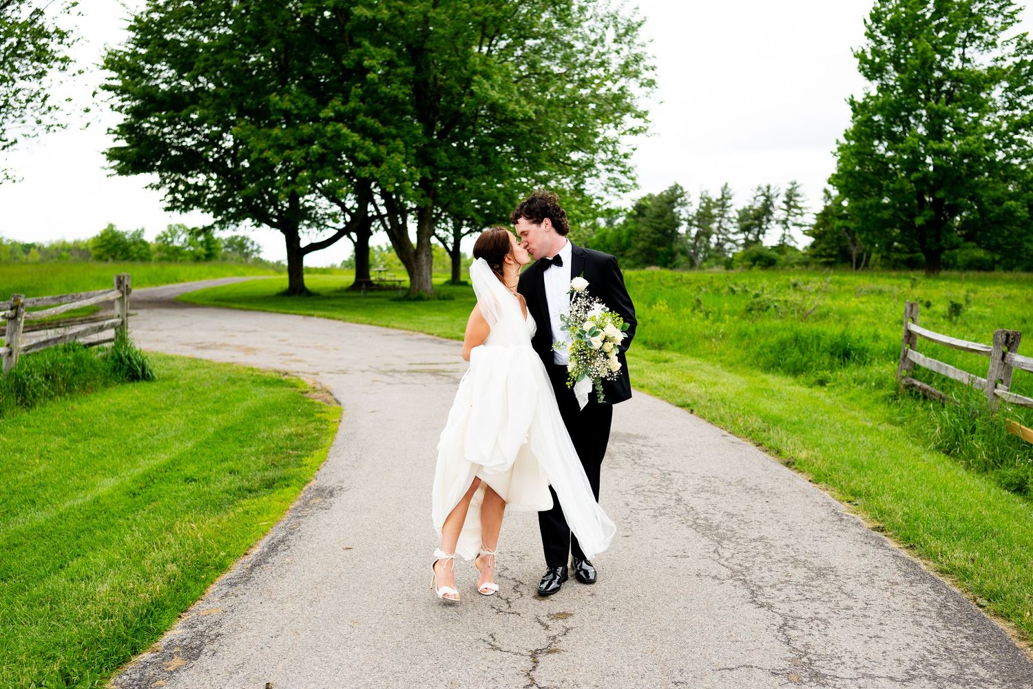 A couple in formal attire shares a romantic moment on a winding gravel path surrounded by lush green scenery.