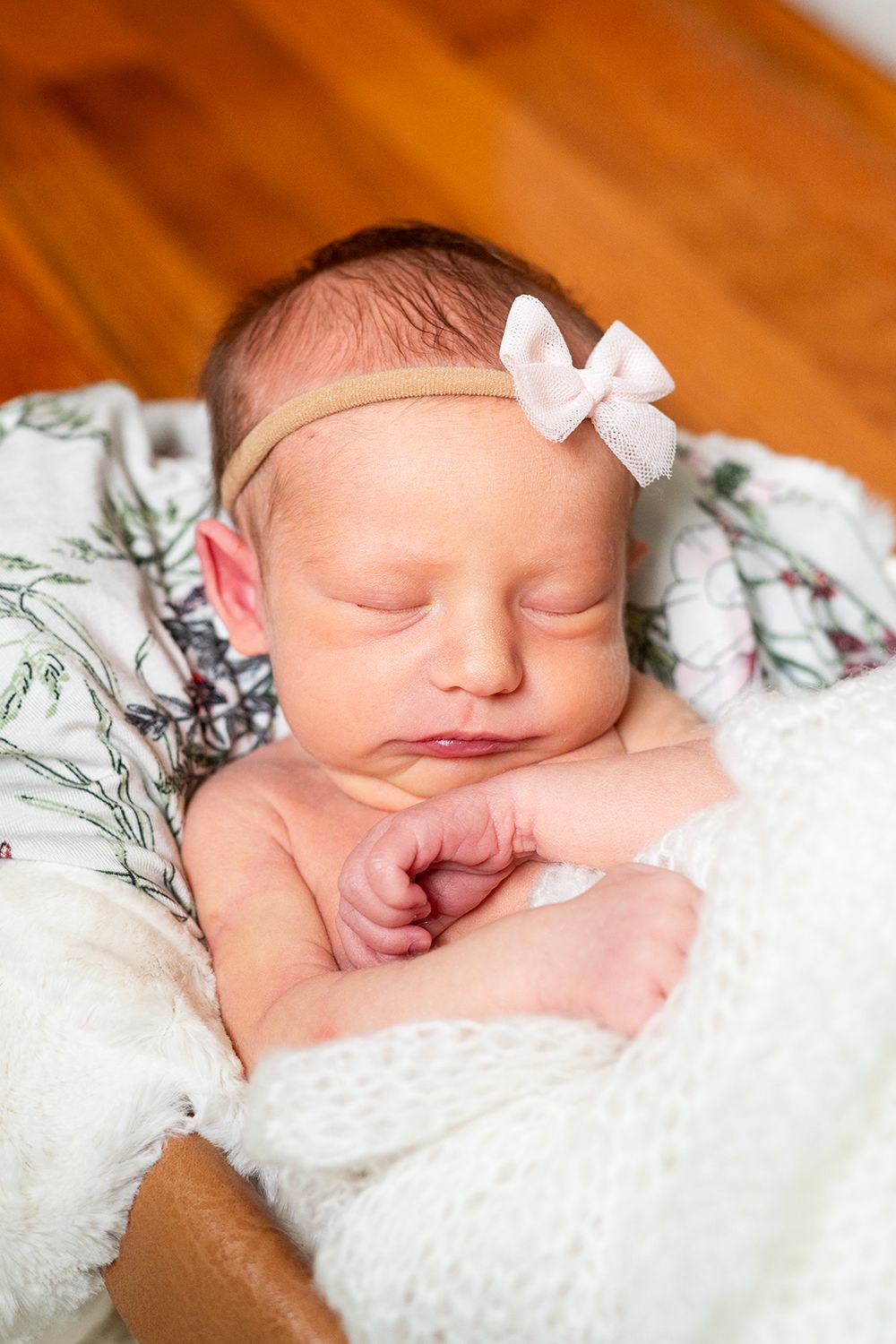 Sleeping newborn baby wearing white bow headband rests peacefully on white blanket against wooden floor.