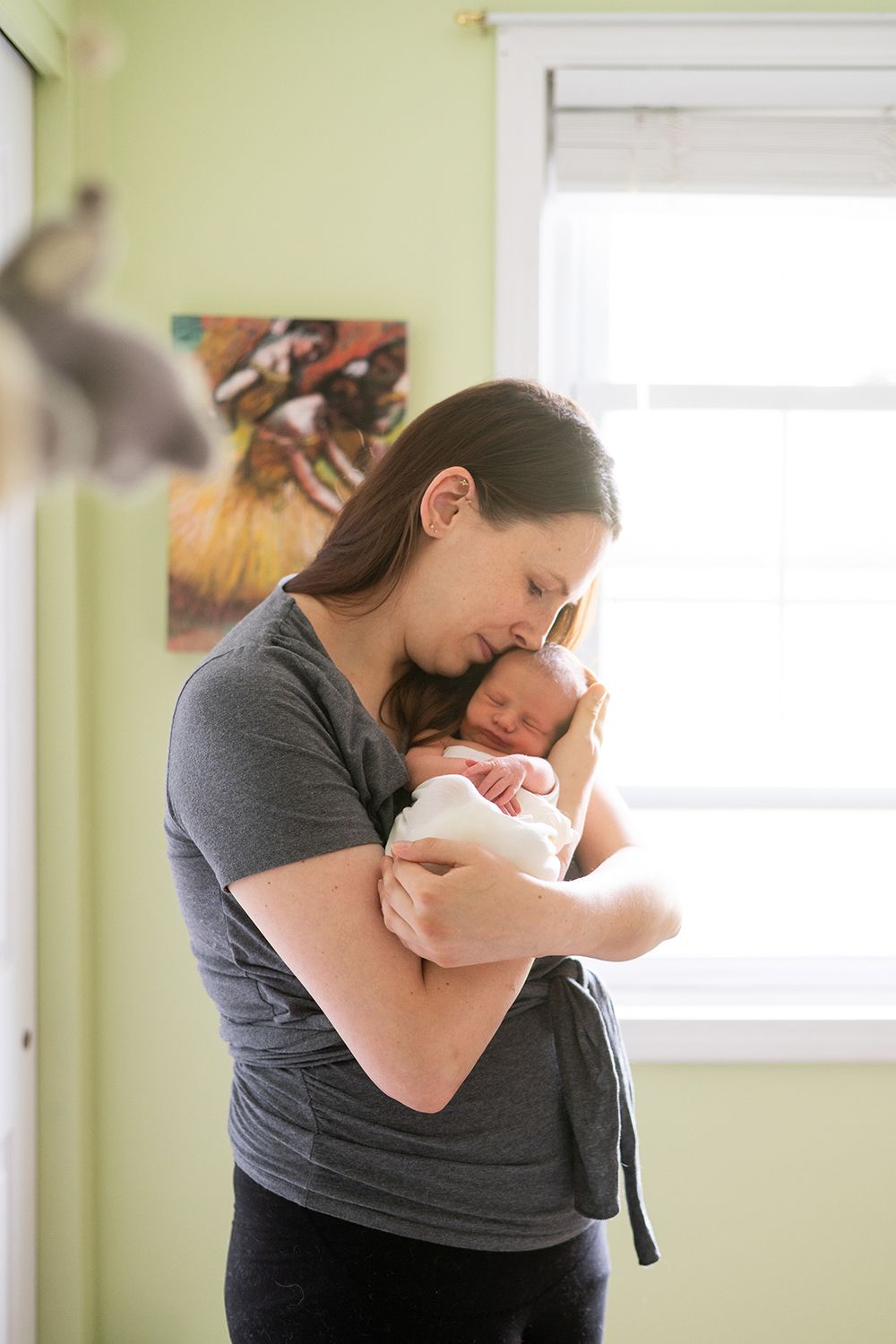 Parent cradling sleeping newborn baby near window in green nursery room.