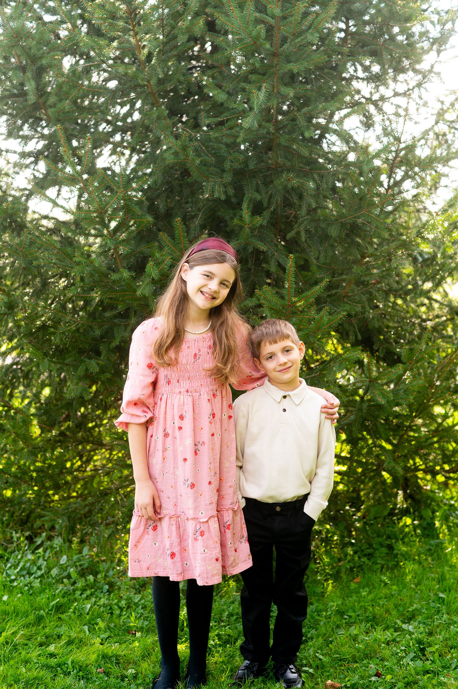 A smiling girl in a pink dress and a young boy in a white shirt pose together outdoors near a green tree.