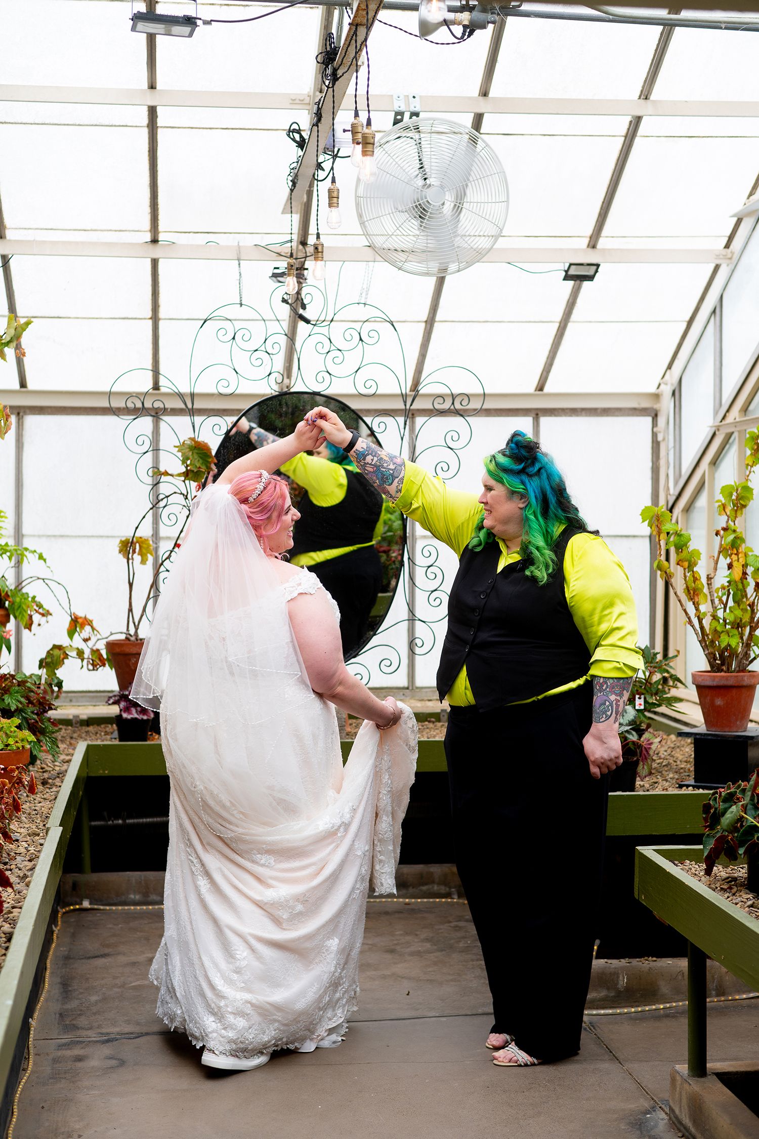 Two brides with colorful hair dance together in a greenhouse, one wearing a white wedding dress and veil.
