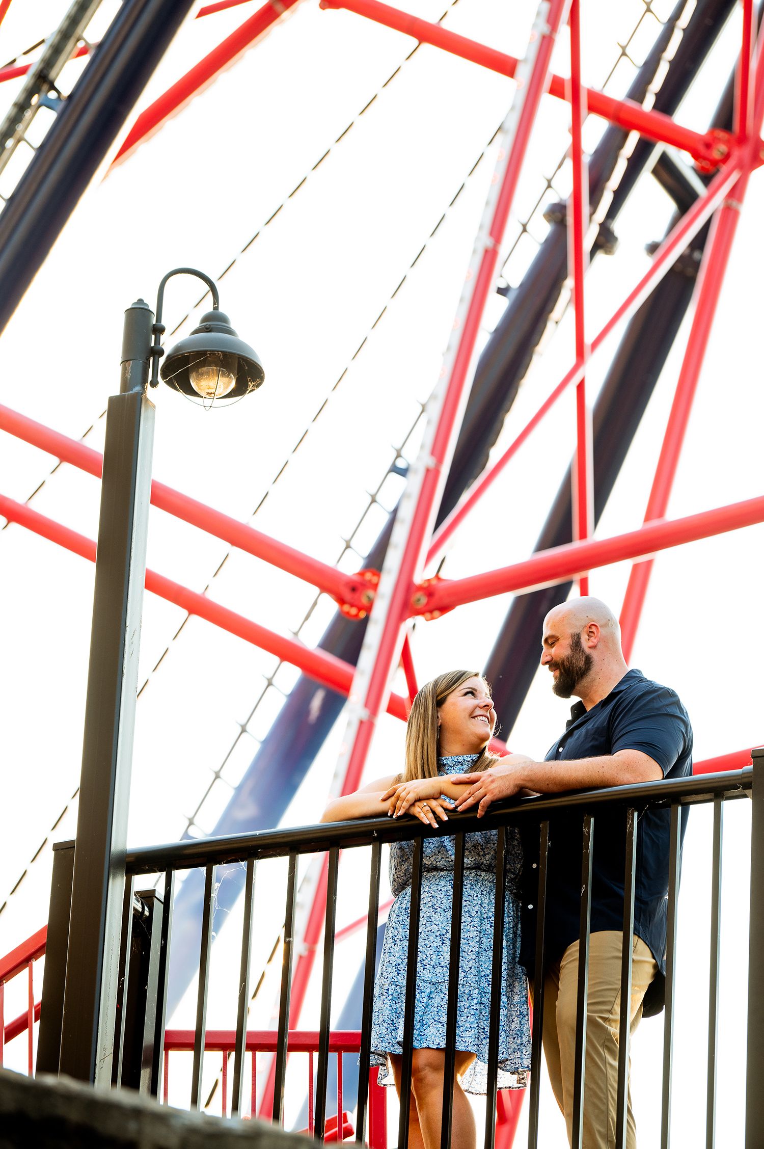 Couple smiling at each other on a balcony near a red Ferris wheel structure and vintage street lamp.
