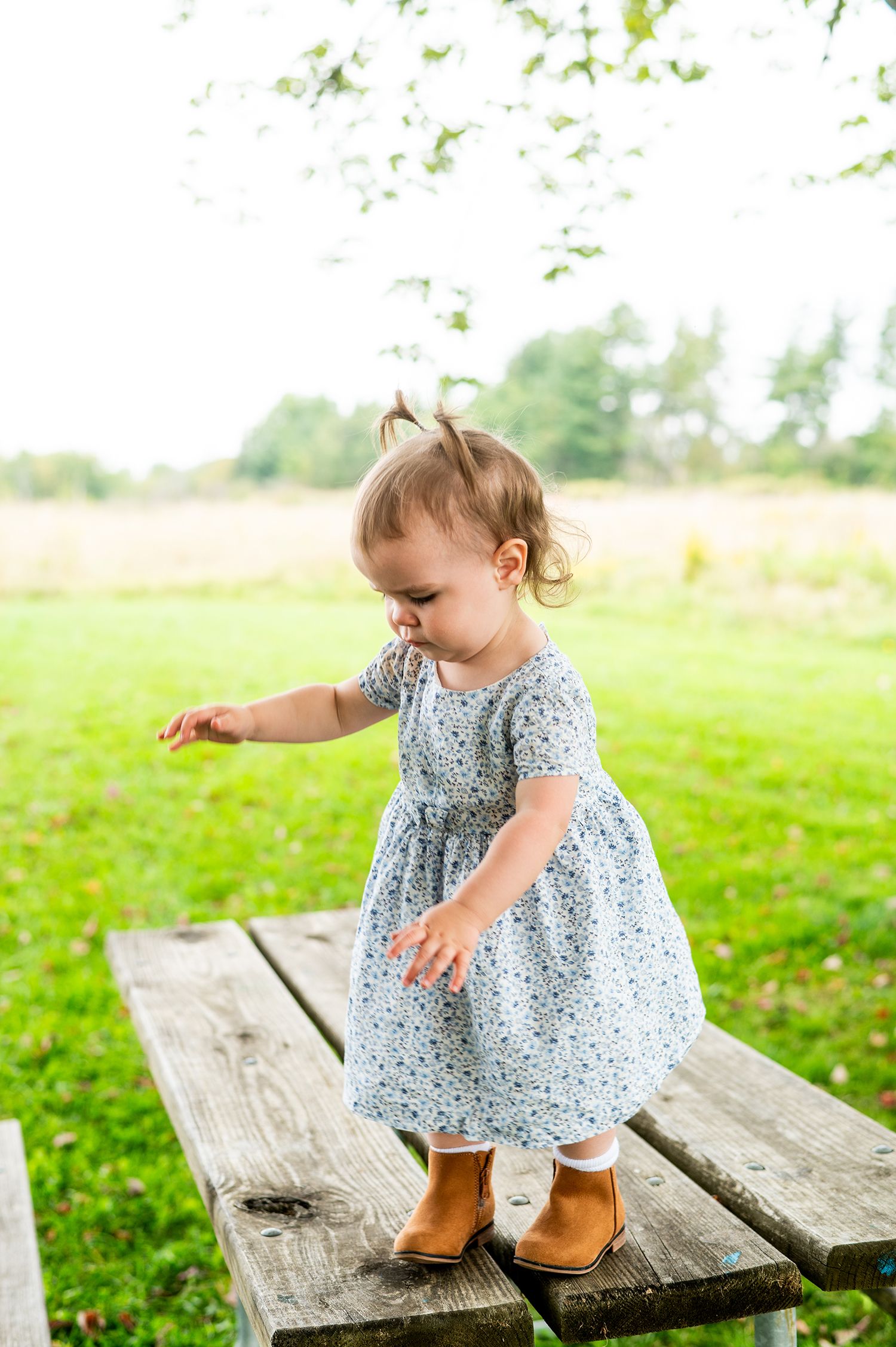 Toddler girl in blue floral dress and brown boots standing on wooden picnic table in green park.