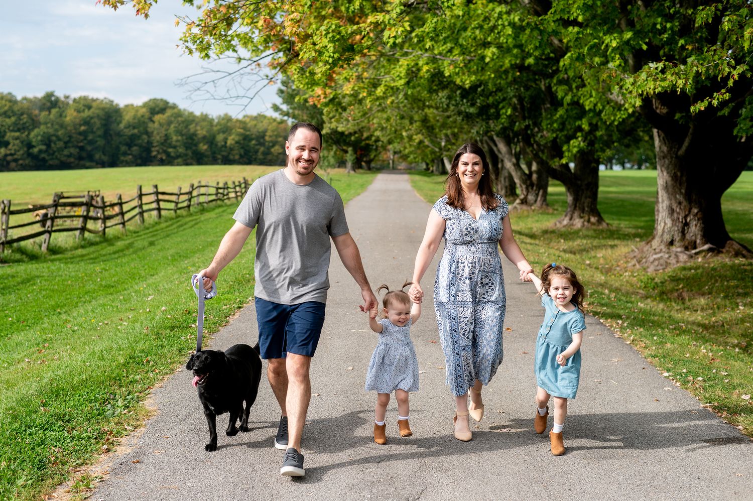 Family of four walking dog on scenic tree-lined path with wooden fence and green fields on a sunny day.