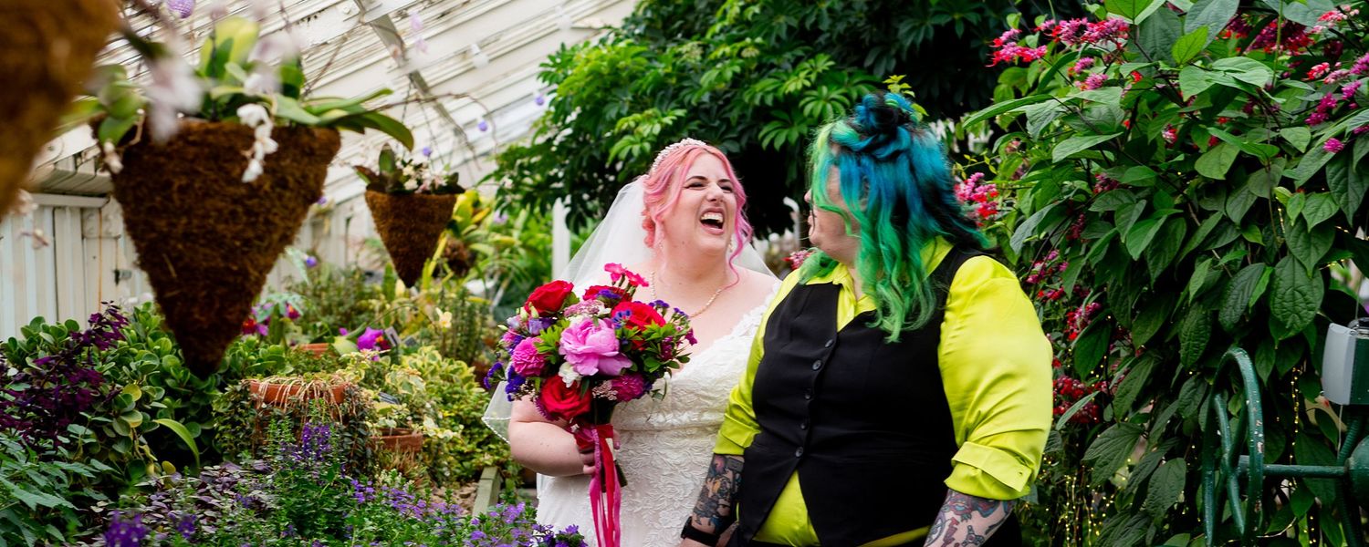Two brides laughing together in a lush greenhouse, one holding a vibrant pink bouquet, wearing wedding attire.