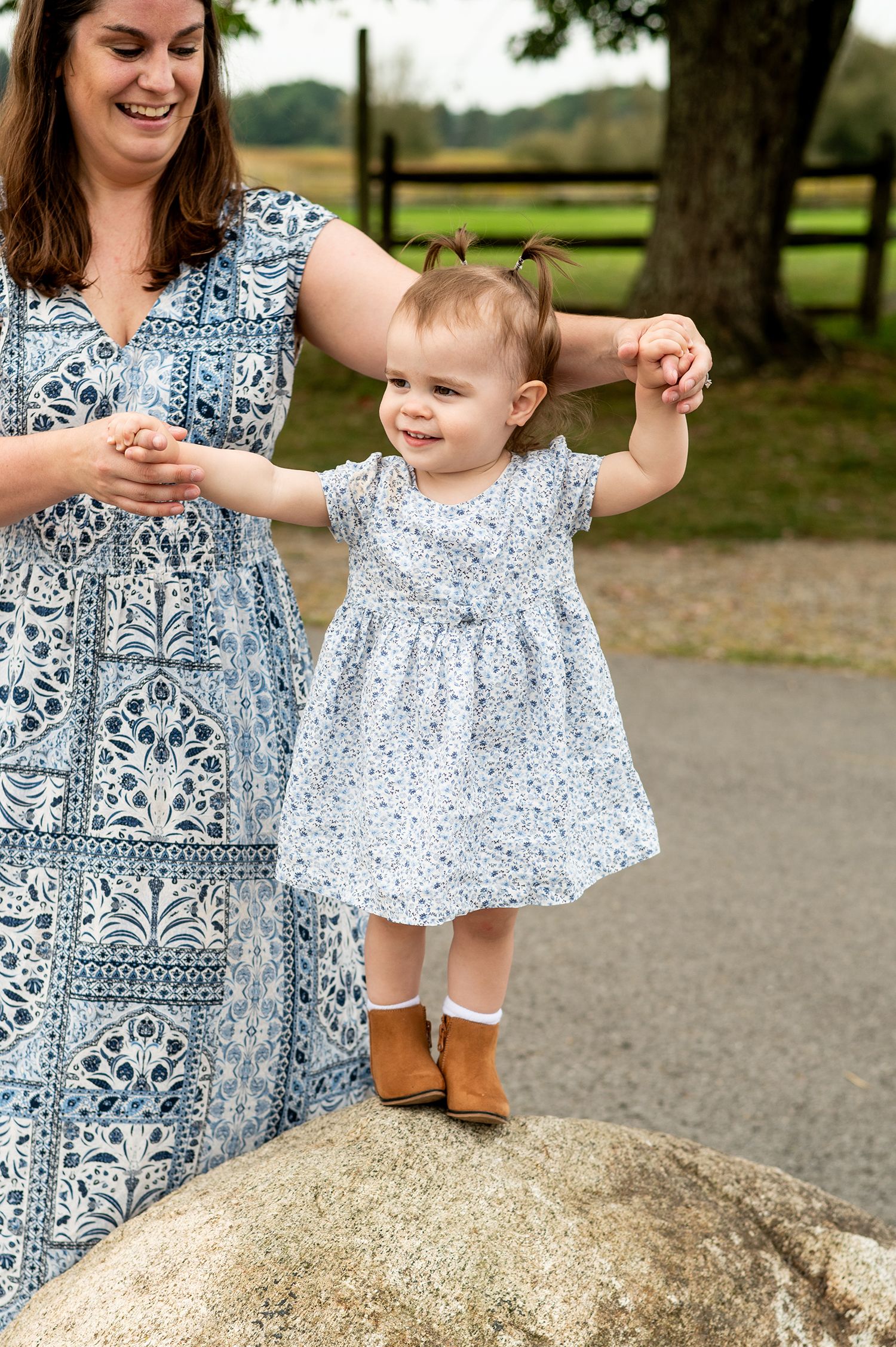 A smiling toddler girl in a white floral dress stands on a rock, held by her mother in a blue patterned maxi dress outdoors.
