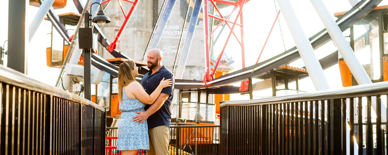 Couple embracing romantically indoors near a colorful Ferris wheel structure at an amusement park.