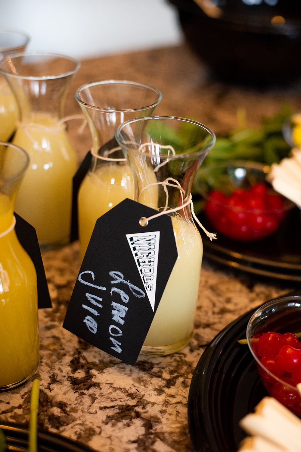Glass bottles of fresh orange juice with black chalkboard tags on a marble countertop next to red garnishes and herbs.