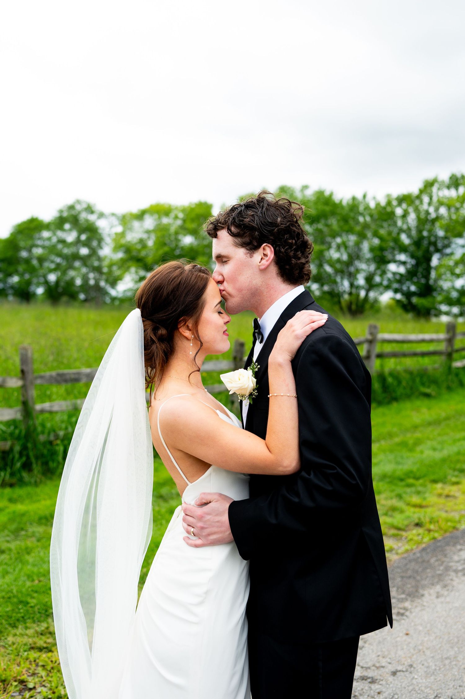 A romantic wedding couple shares a tender kiss on a grassy hillside path with trees in the background.