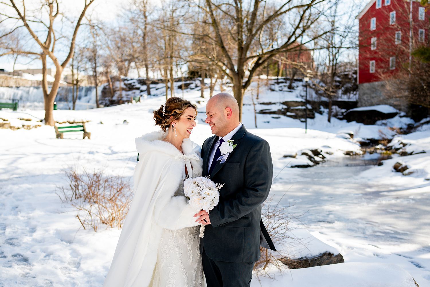 Newlyweds dance surrounded by snow-covered grounds and a red building visible in the distance.