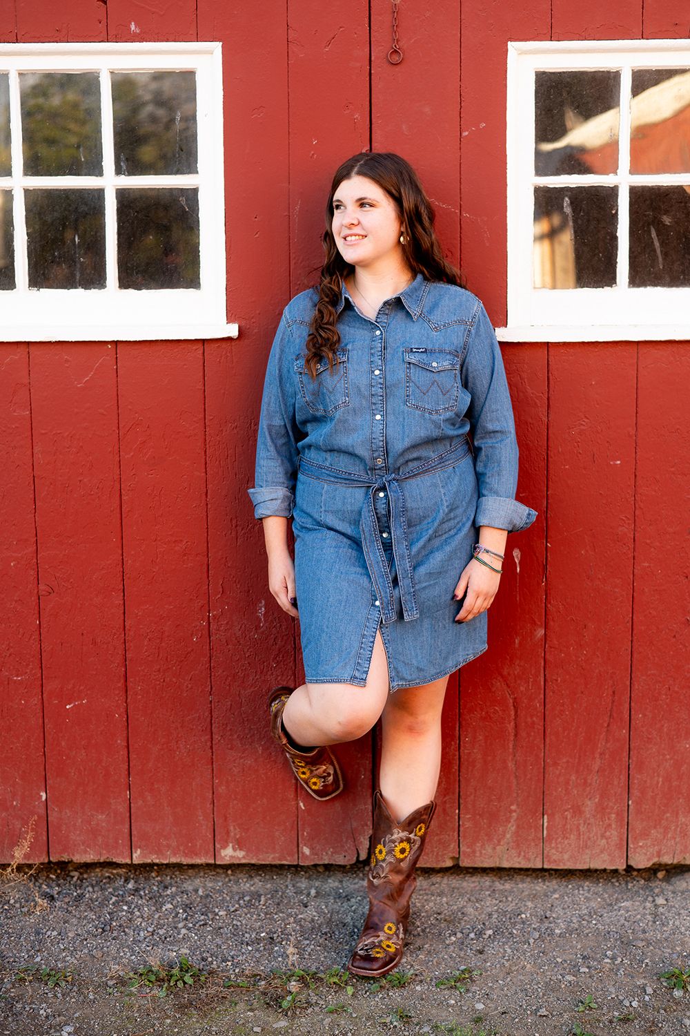 A person in a denim shirt and brown boots leans casually against a red barn wall with white-trimmed windows.