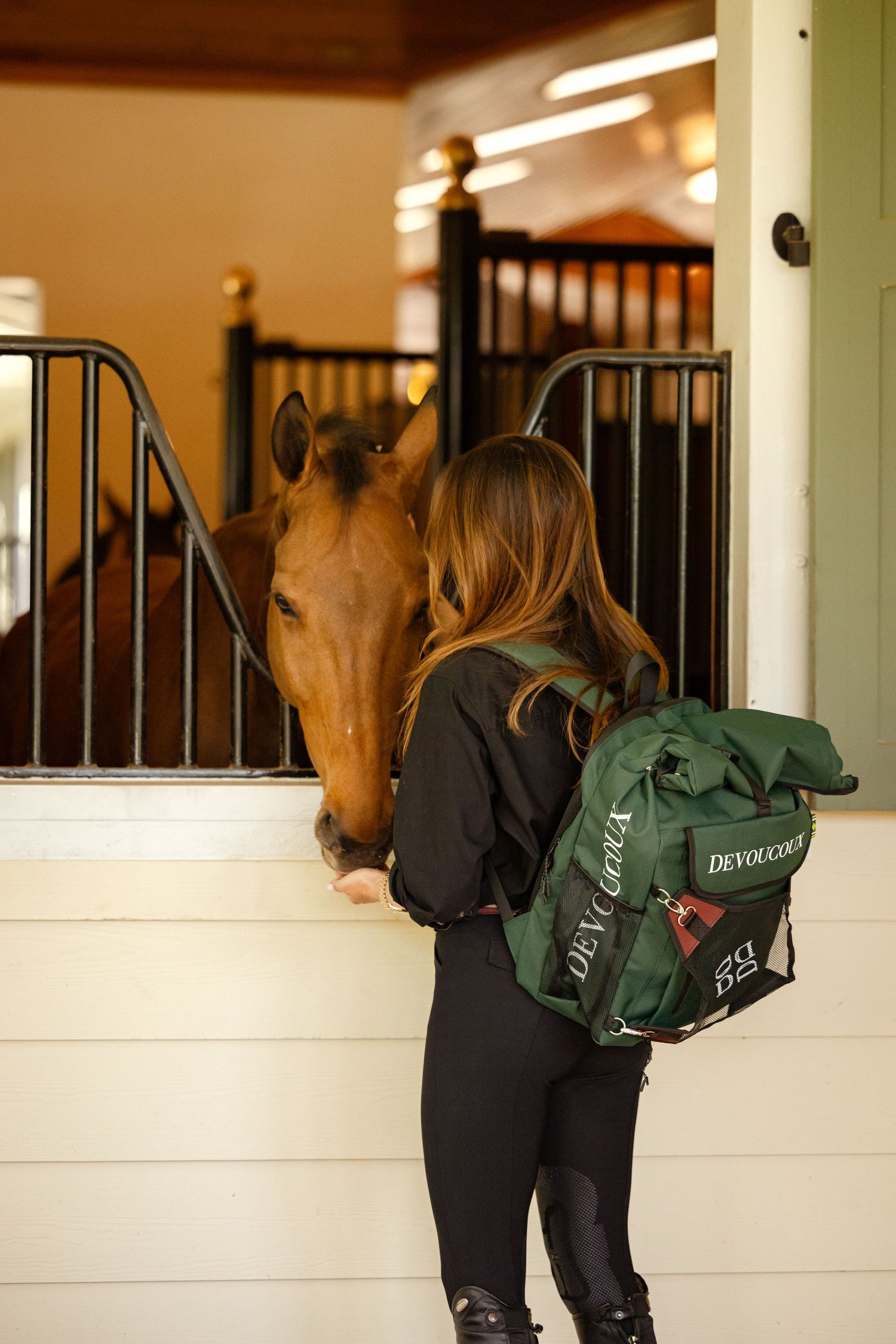 Rider with green backpack interacting with horse in stable stall.