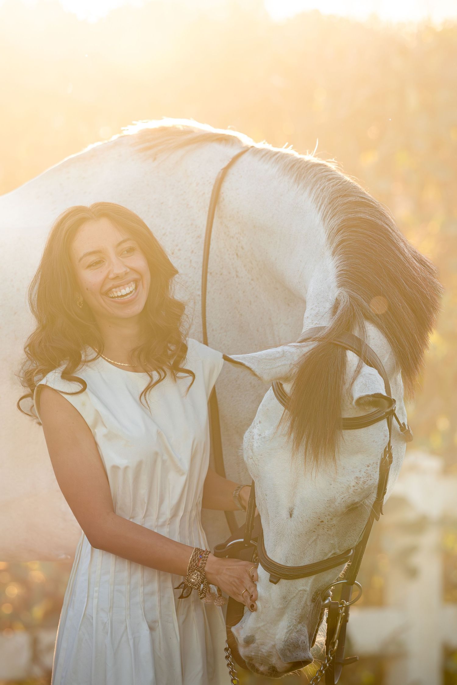 A person in a white dress shares joyful moments with a white horse during a golden sunset photoshoot.