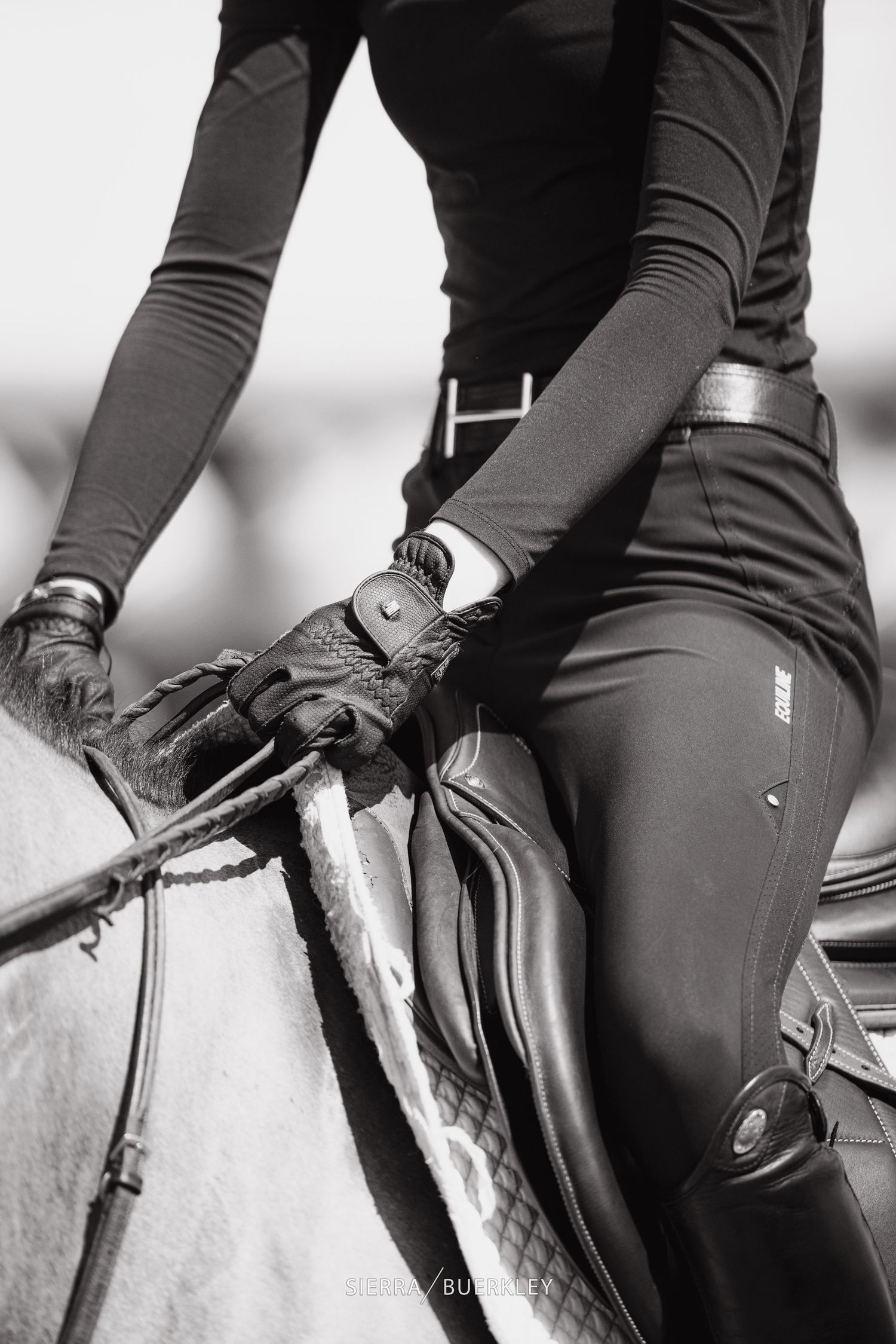 Close-up black and white photo showing rider in equestrian gear with riding gloves holding leather reins on horseback.