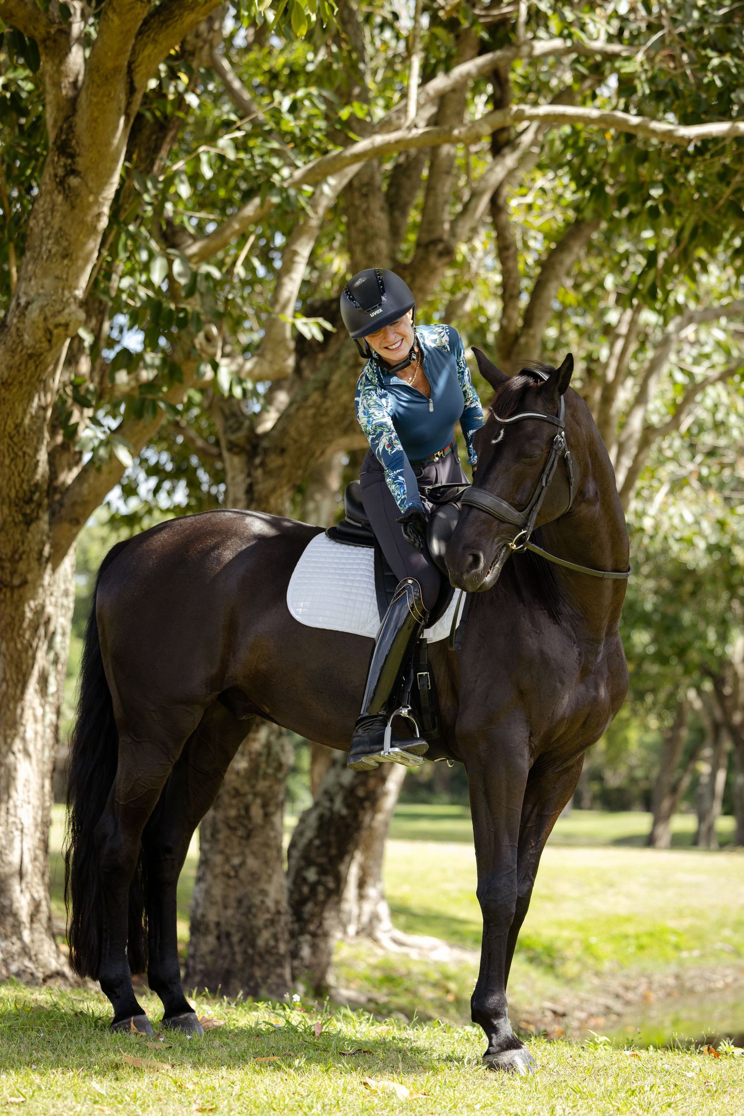 Equestrian rider in dark clothing sits atop a majestic black horse in a sunny wooded park setting.