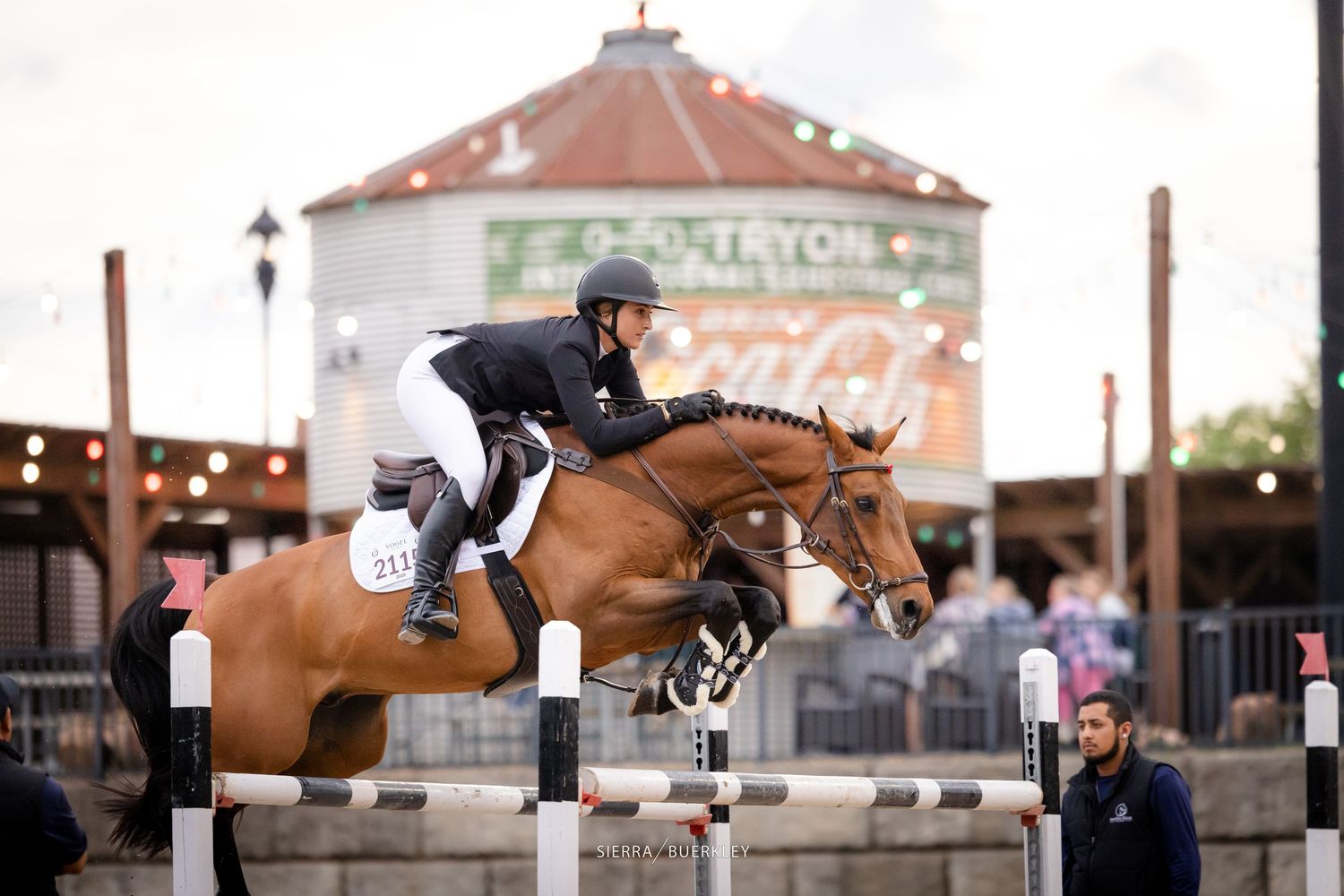 Equestrian rider and brown horse jumping over obstacle at competition with farm building in background.