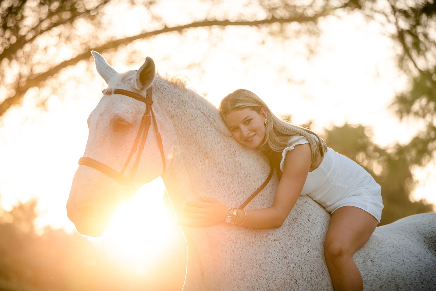 A white horse and rider pose in a golden sunset with lens flare creating a dreamy backlit effect.