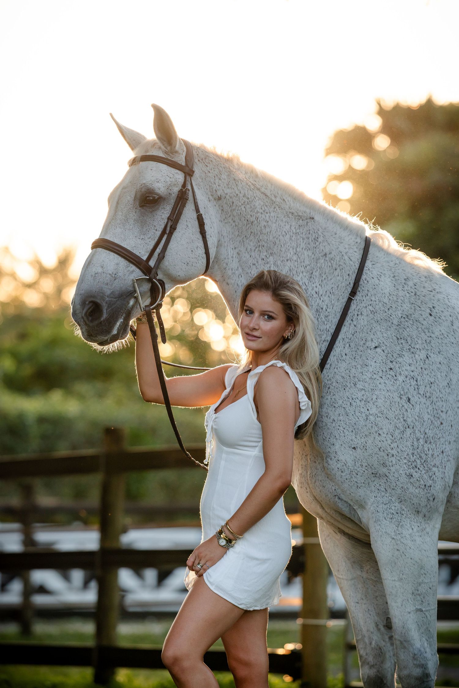 A white horse and rider pose together at sunset against a rustic wooden fence in an equestrian photoshoot.