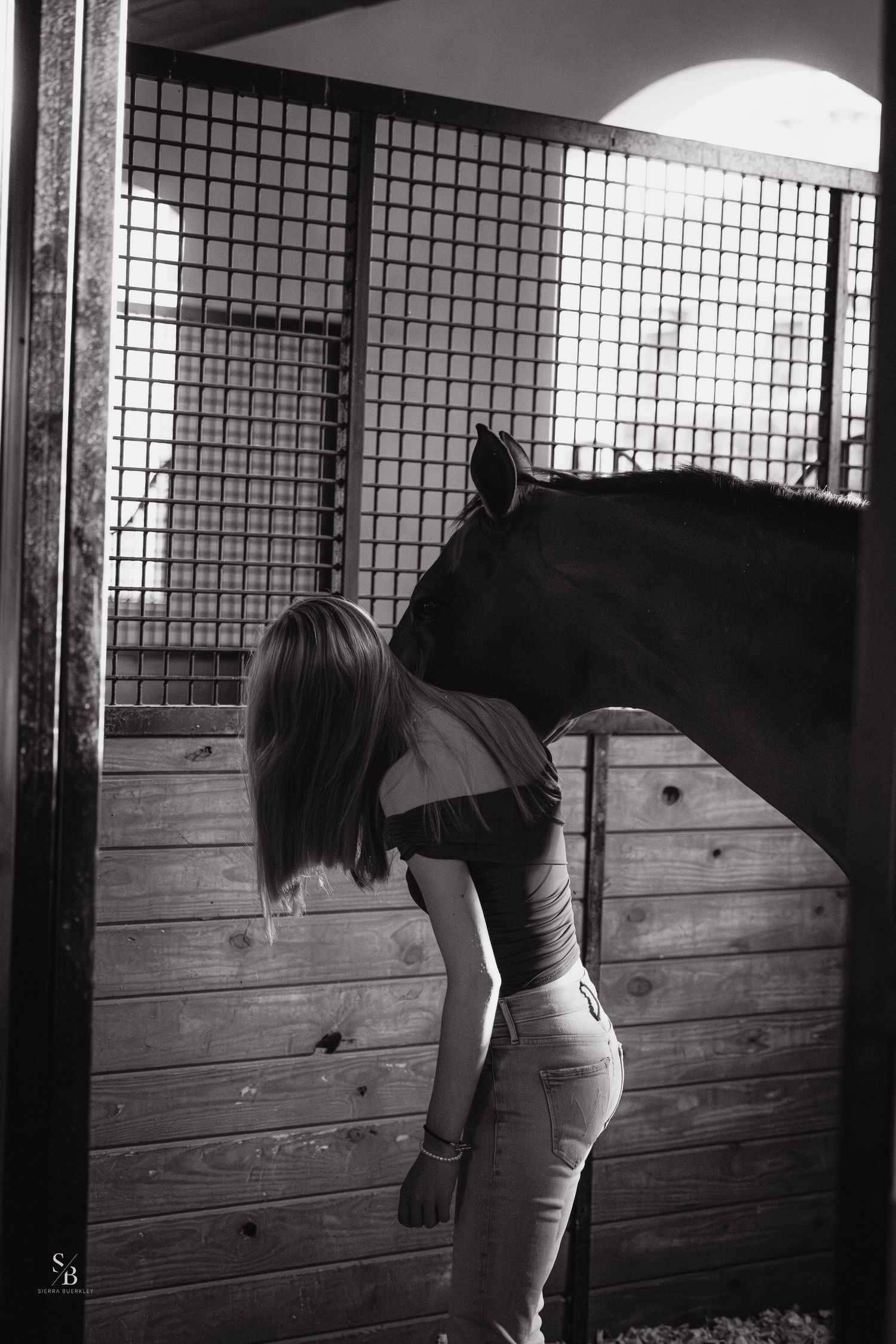 Black and white photo of a person bonding with a dark horse in a wooden stable.