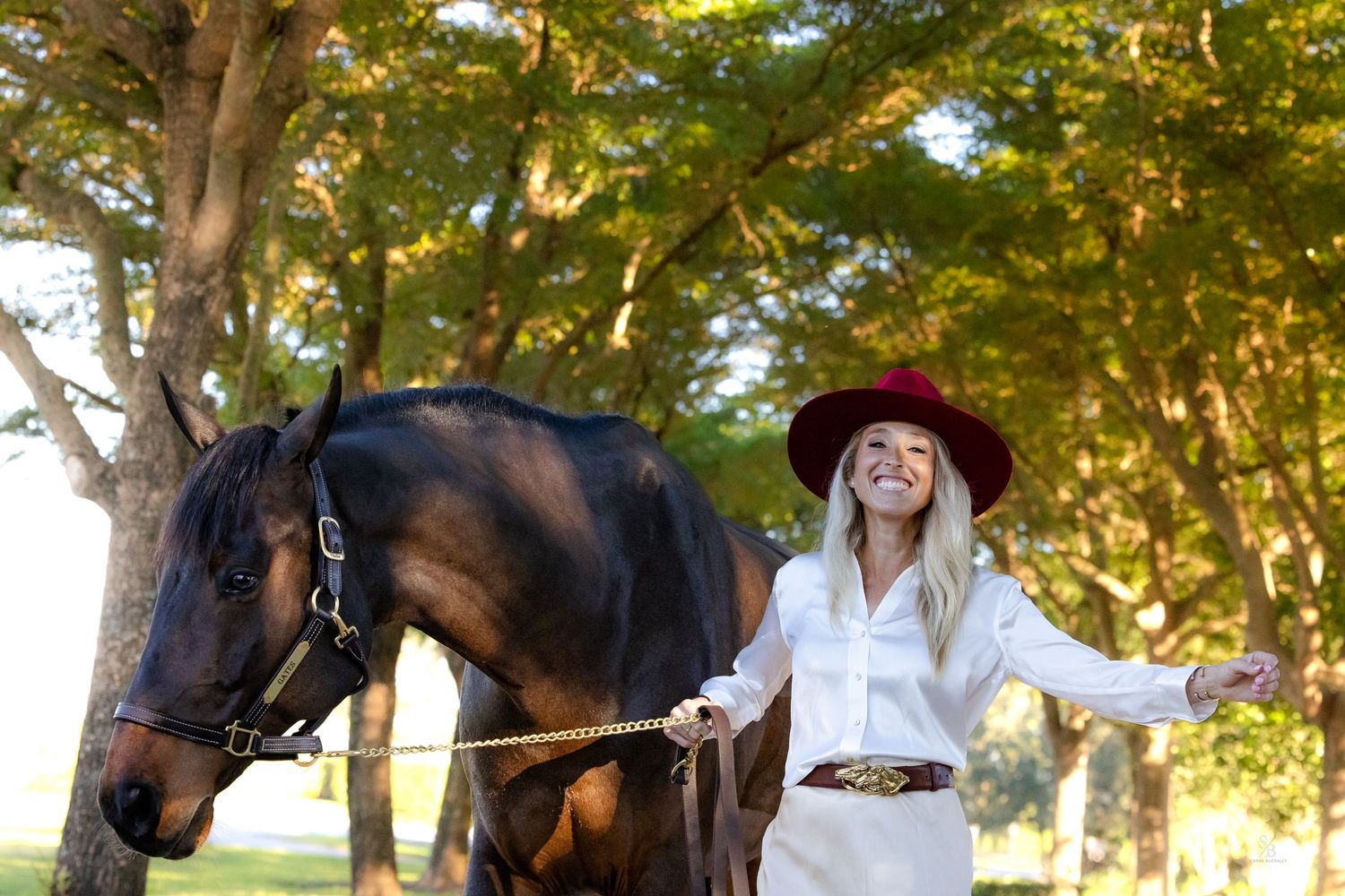 Person in western attire and red hat poses with black horse under autumn trees.