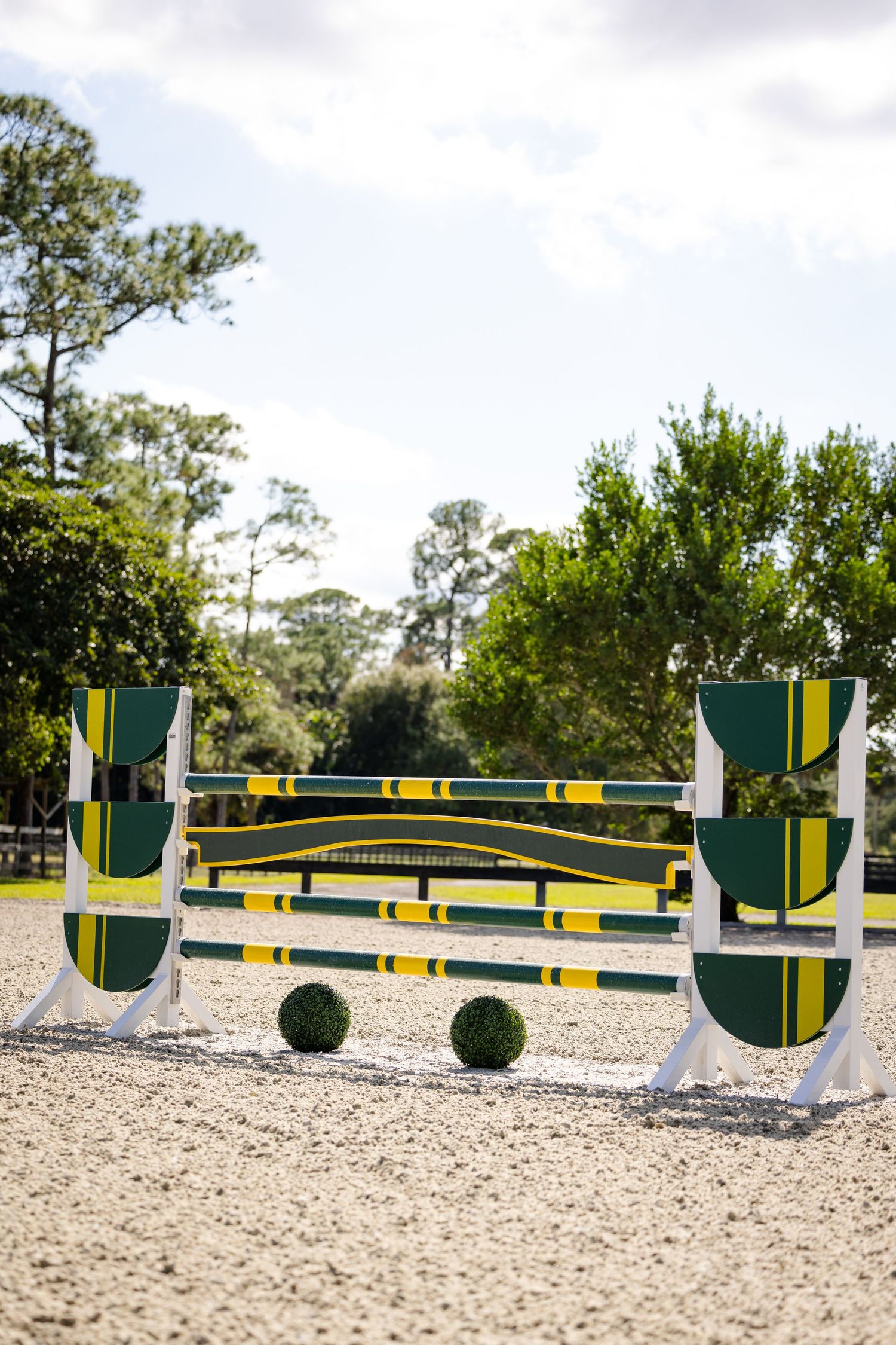A green and yellow striped horse jumping obstacle set up in a sandy arena with pine trees in the background.