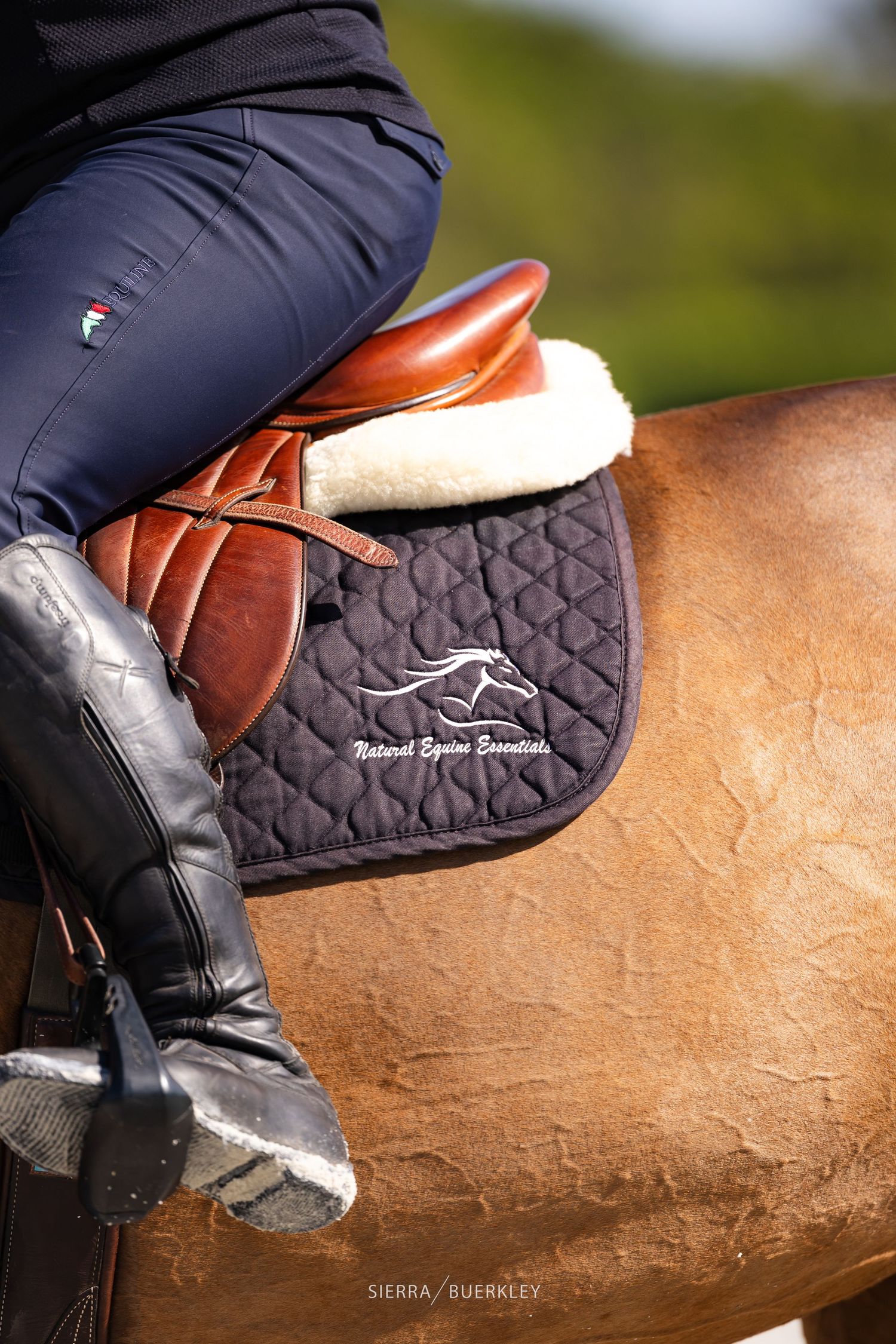 Close-up view of equestrian gear showing leather saddle, black pad with embroidered logo, and riding boots on horseback.