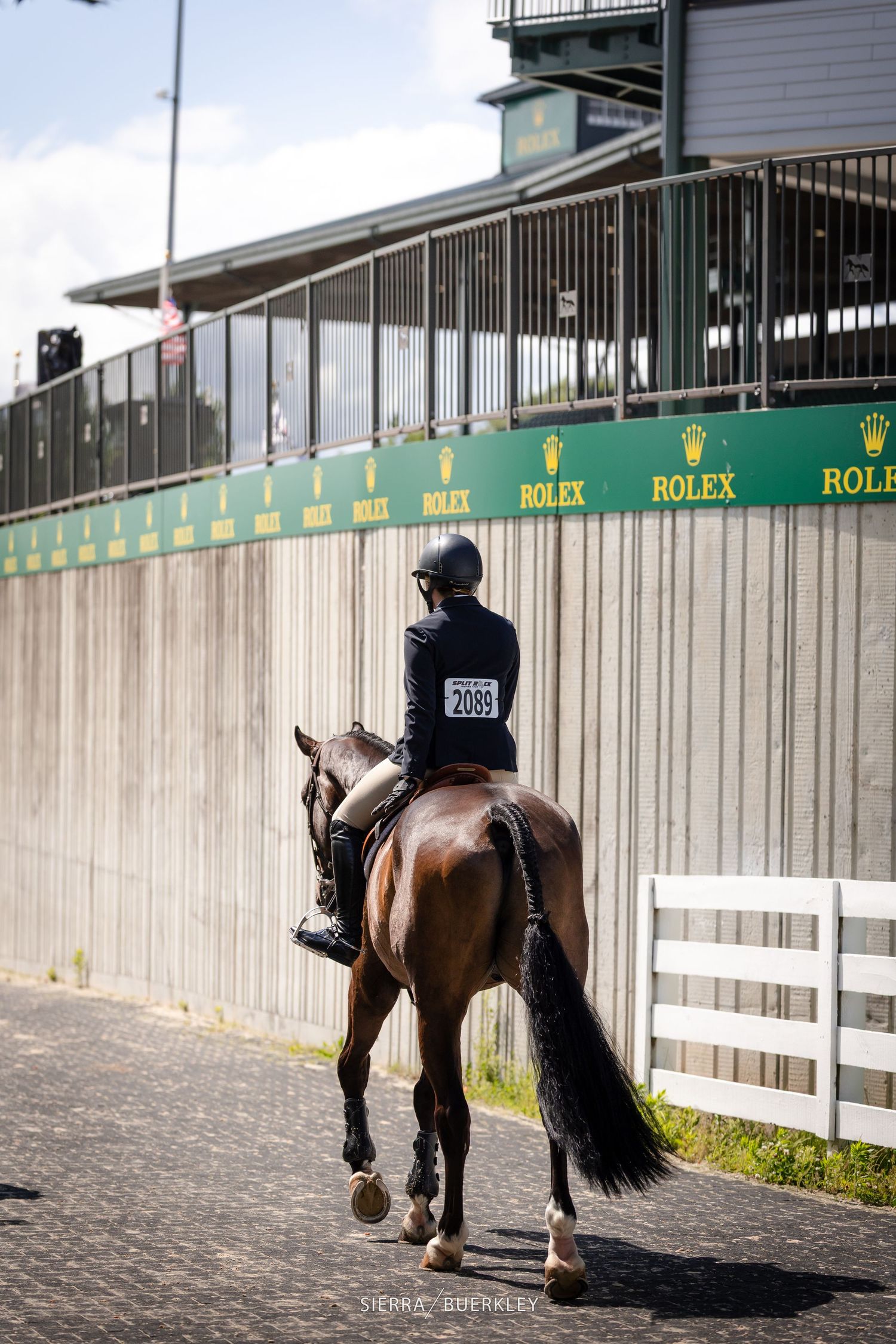 An equestrian rider wearing a black jacket and helmet rides a brown horse past Rolex advertising banners at an event.