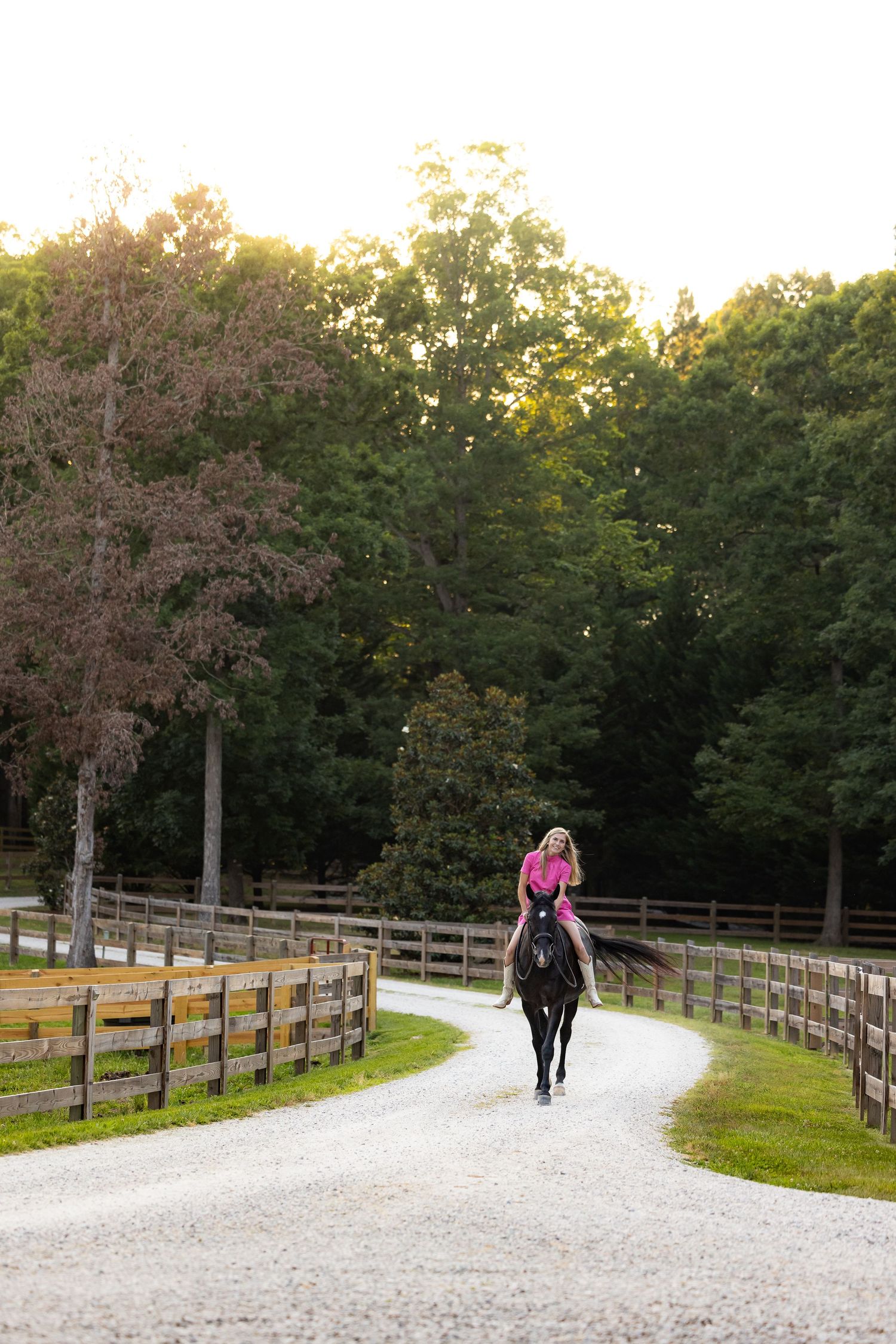 A rider guides their horse along a winding gravel path bordered by wooden fences at sunset.