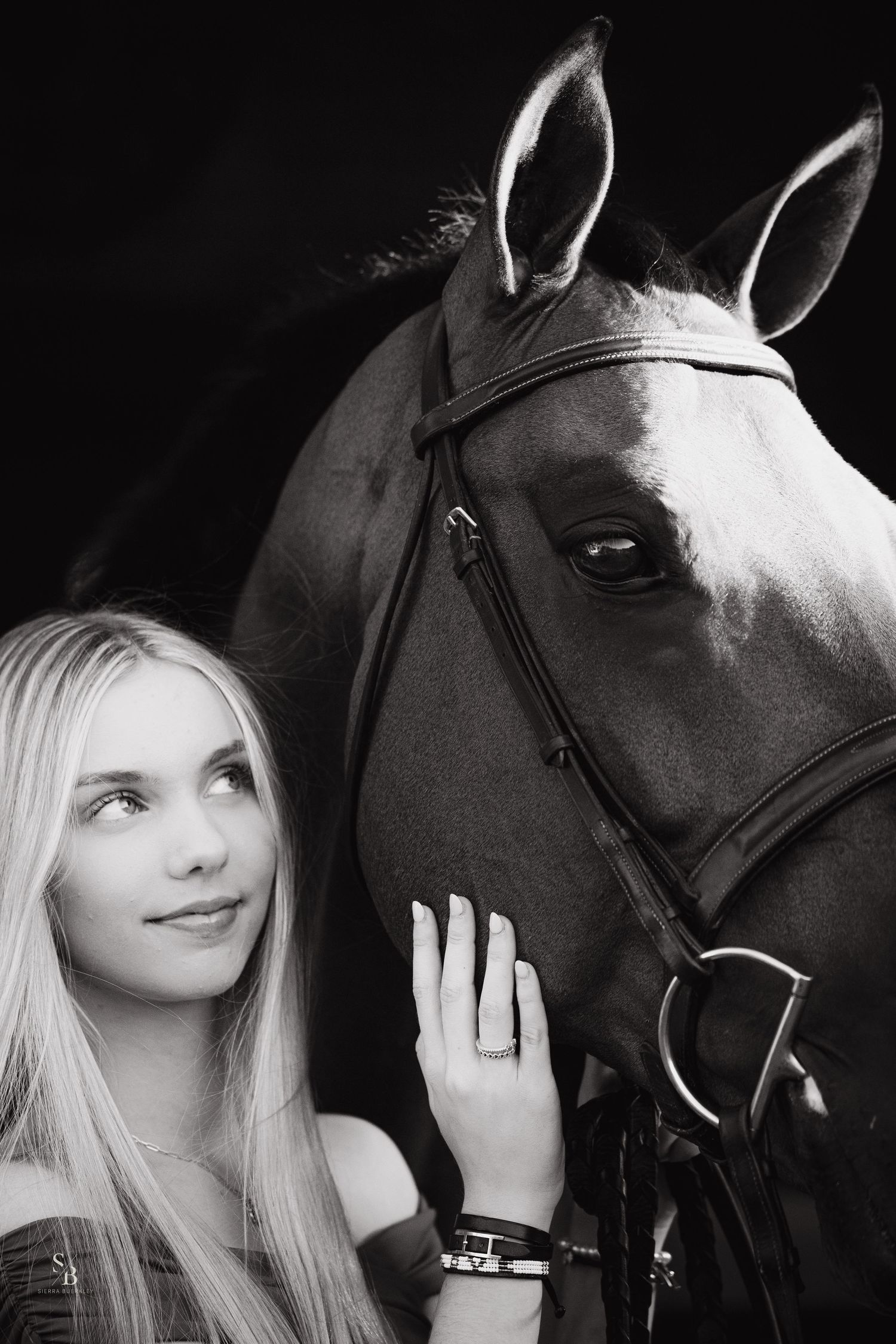 A black and white photograph of a person posing with a dark horse wearing bridle and reins.