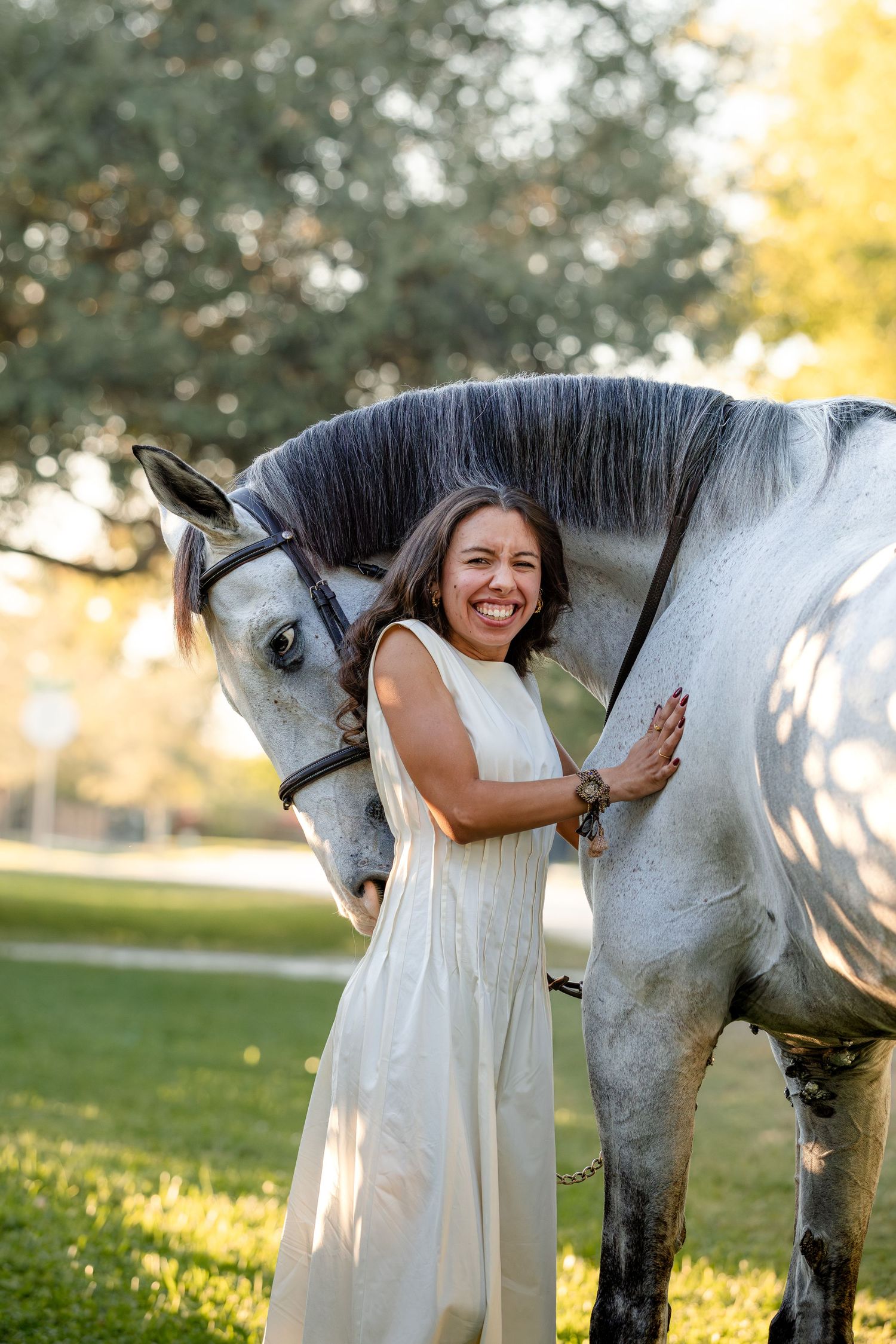 A woman in a white dress hugs and interacts with a beautiful white horse in a sunlit grassy field.