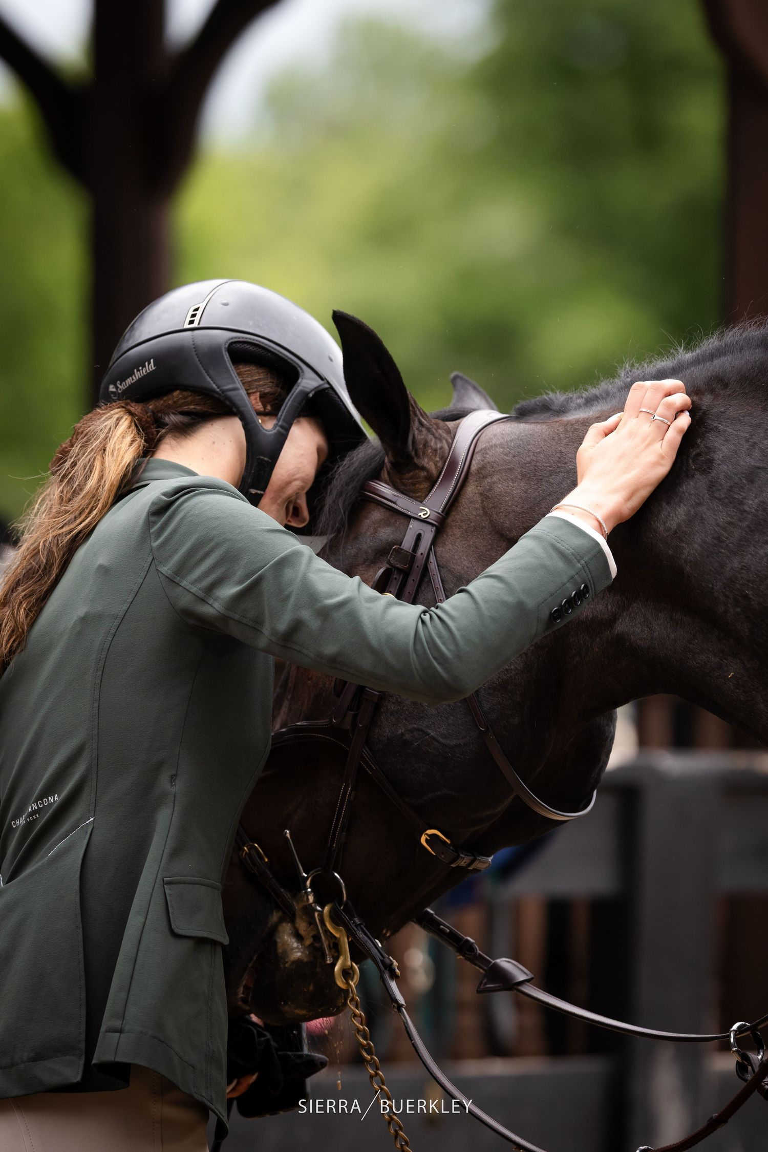Equestrian rider in green show coat bends over to adjust the saddle on a dark horse.