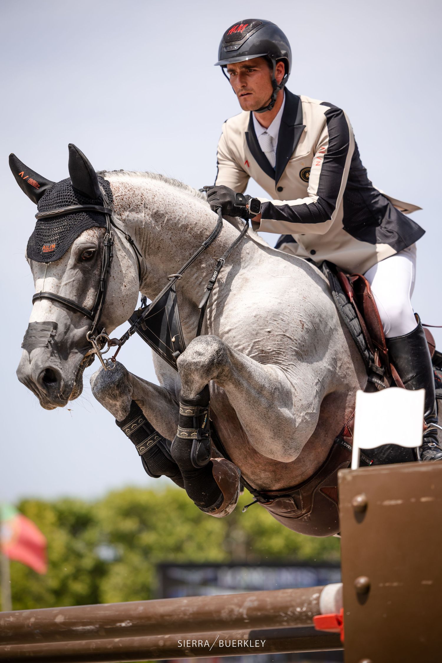 Equestrian rider in white and black attire jumps a grey horse over show jumping obstacle during competition.