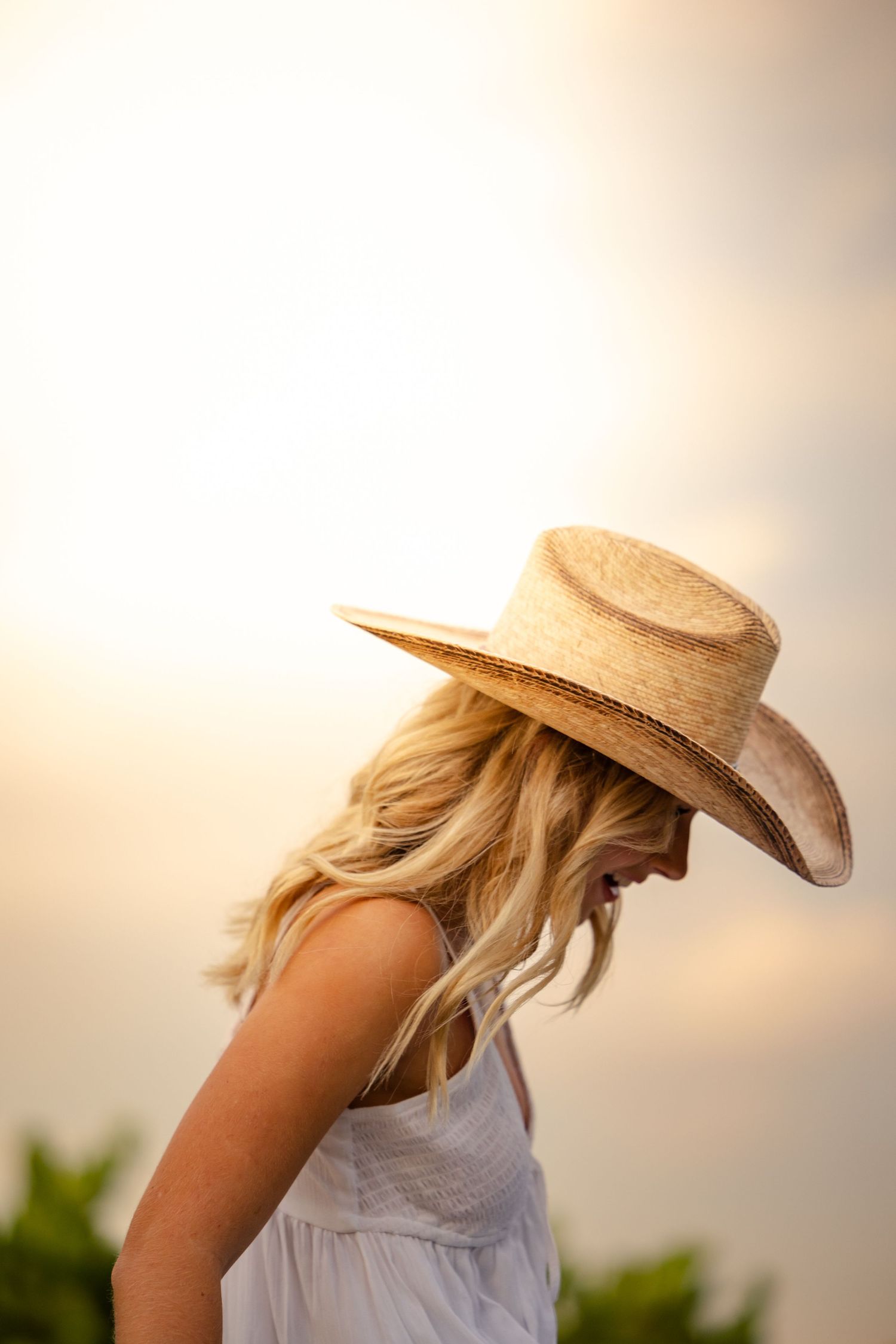 A person in a white dress wearing a straw cowboy hat stands against a soft sunset backdrop.